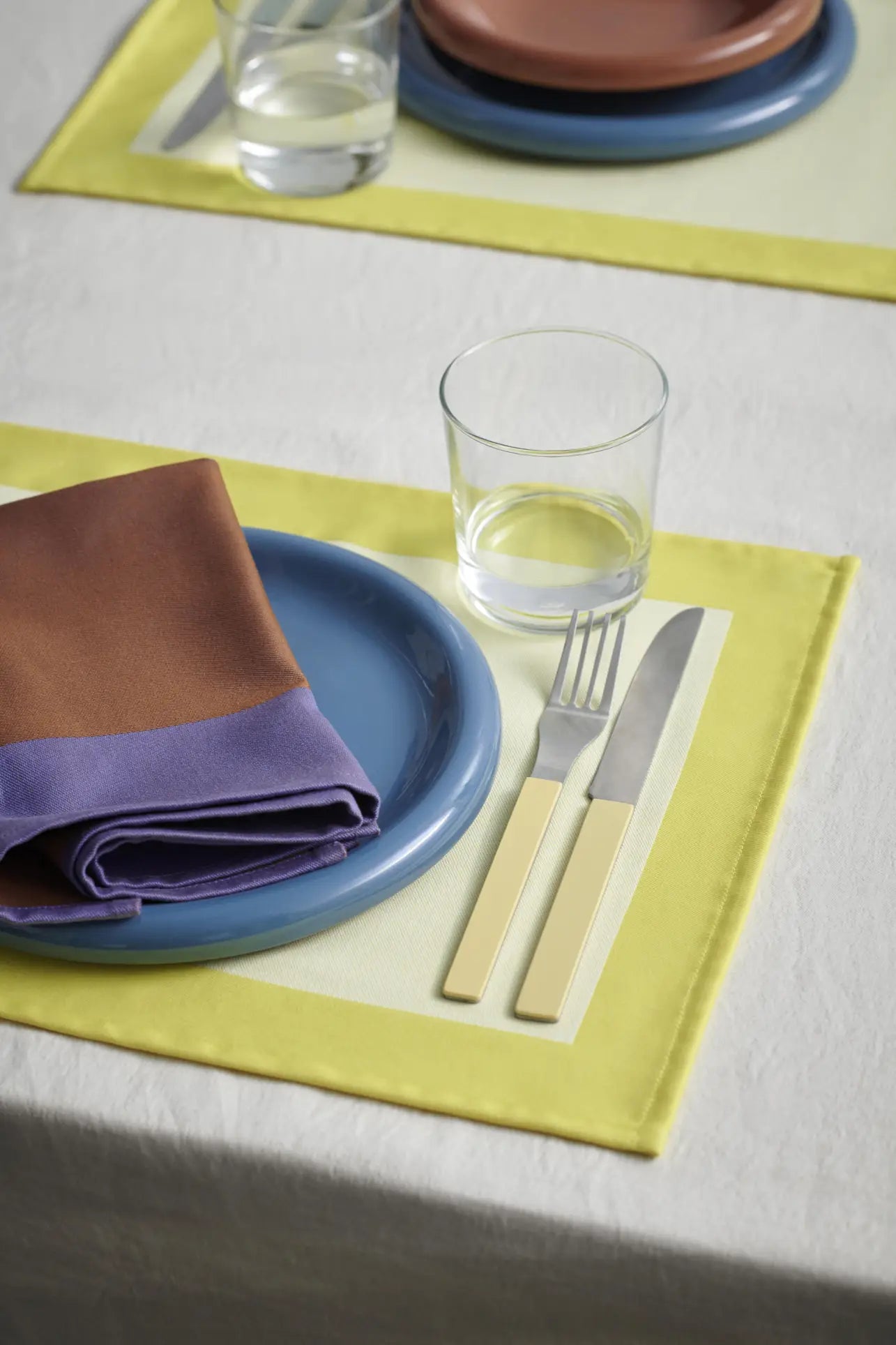 A tableware setting features the HAY Barro Plate 24cm, Set of 2 by HAY in blue, paired with a brown and purple napkin, water glass, terracotta-style cream cutlery on a yellow placemat over a white tablecloth.