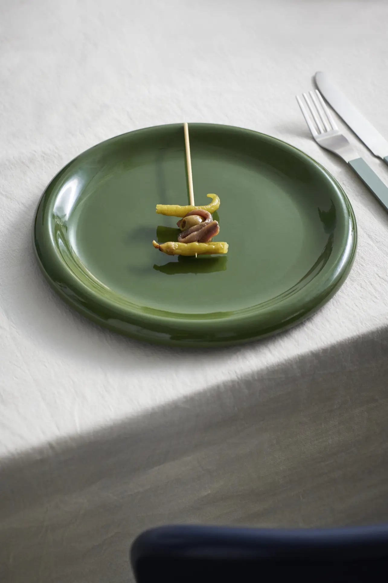 A single skewer sits on a green HAY Barro Plate 24cm, Set of 2, by HAY atop a white tablecloth. A knife and fork from the tableware collection are placed to the right of the plate.