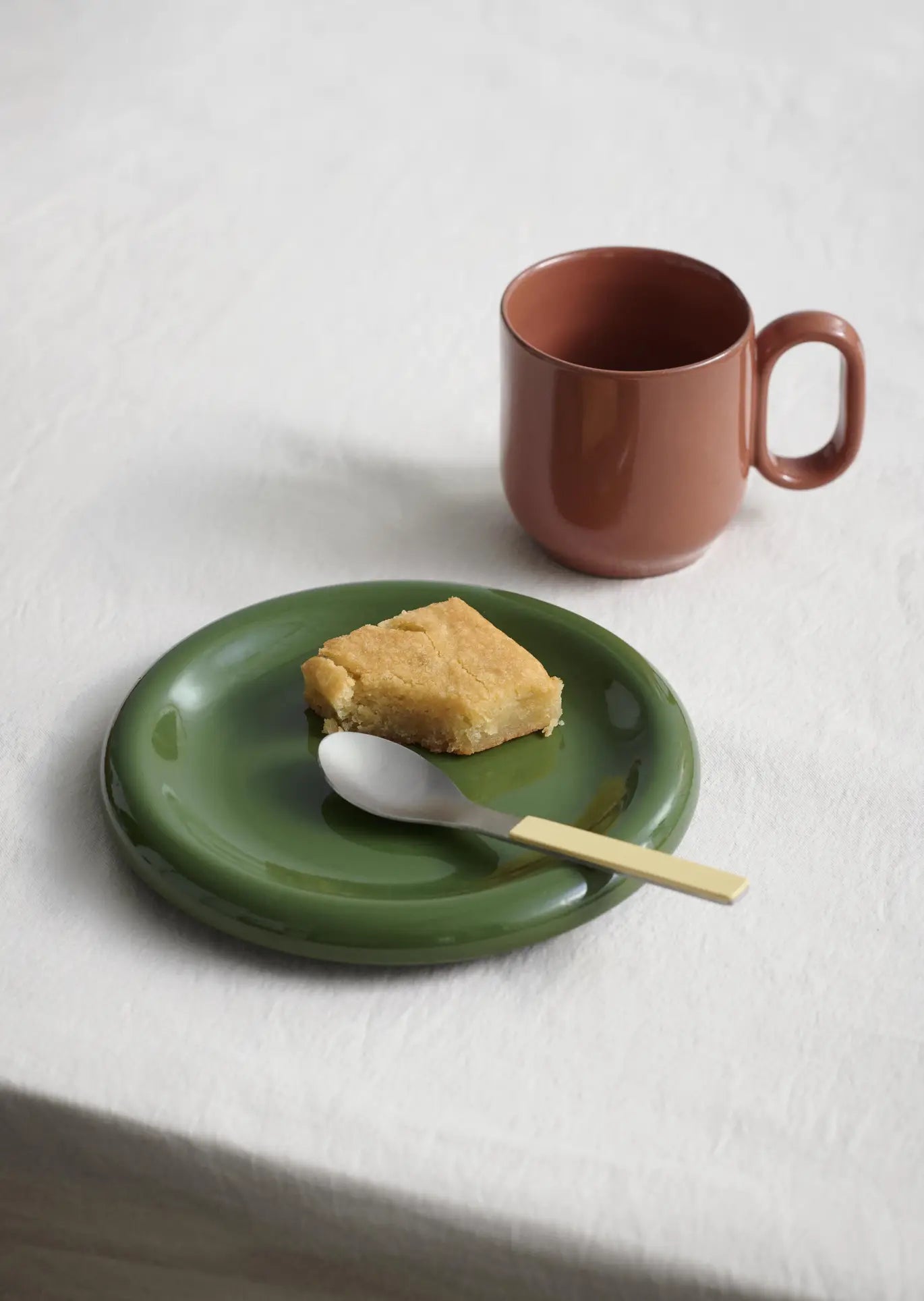 A slice of cake rests on a HAY Barro Plate 24cm from HAY, paired with a spoon and brown mug on a white tablecloth—an inviting glimpse from our curated tableware selection.
