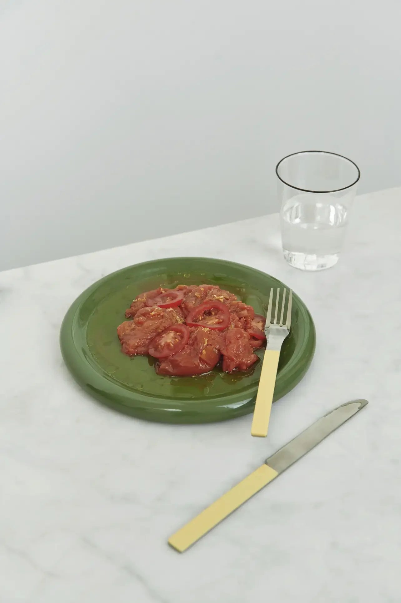 A HAY Barro Plate 24cm from HAY, set of 2, displays sliced tomatoes and sauce with a fork on the plate and a knife nearby on a white marble table. A glass of water completes this elegant tableware scene.