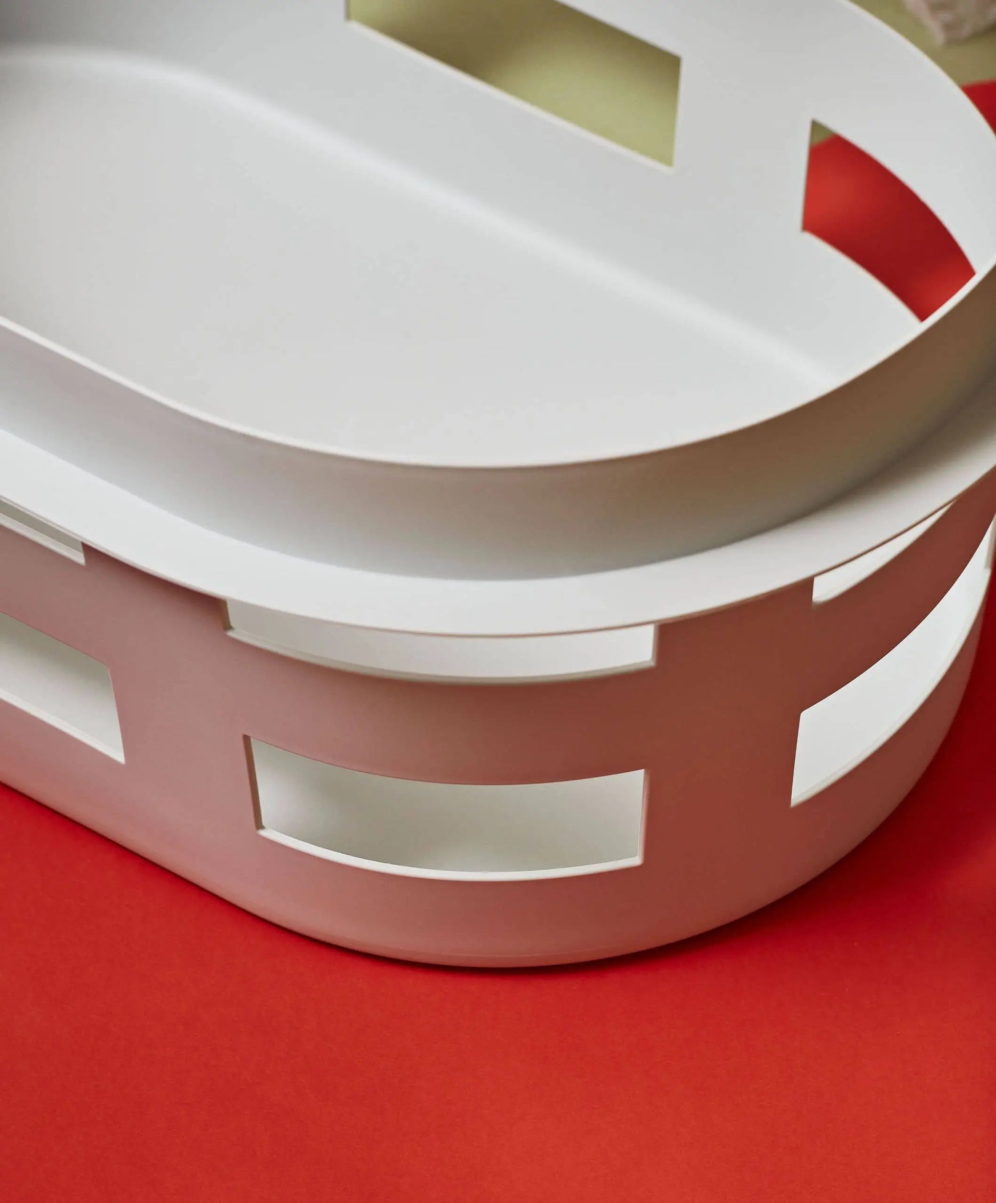 The HAY Basket S by HAY is a modern, white, oval container with rectangular cutouts, shown on a red surface from an overhead angle.