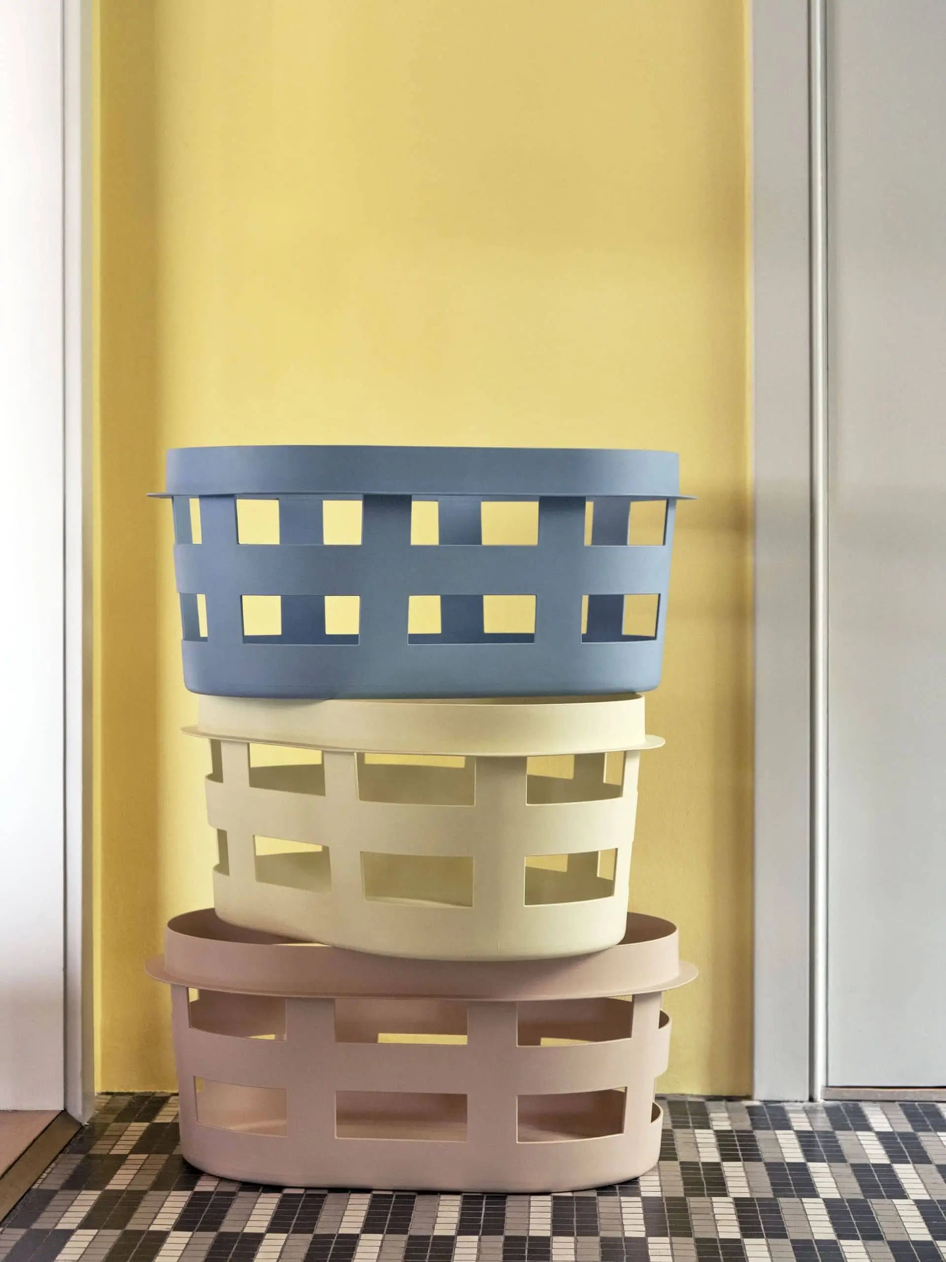 Three HAY Basket S plastic laundry baskets by HAY, in pastel blue, cream, and beige, are stacked together against a yellow wall on a black-and-white patterned tiled floor.