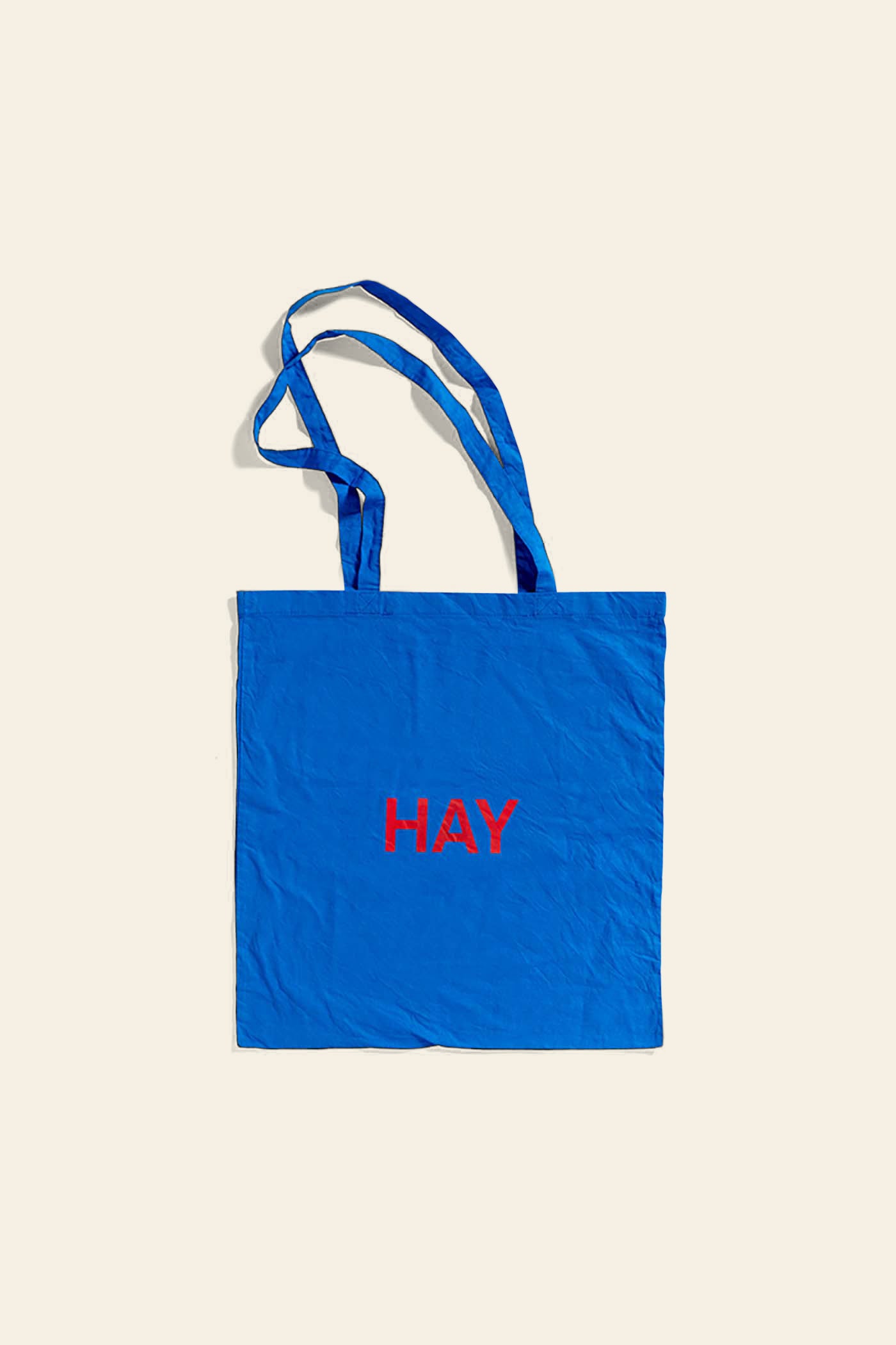 The HAY Blue Tote Bag, Red Logo, by HAY, is a cotton tote with two handles and HAY in red letters on the front, shown against an off-white background.