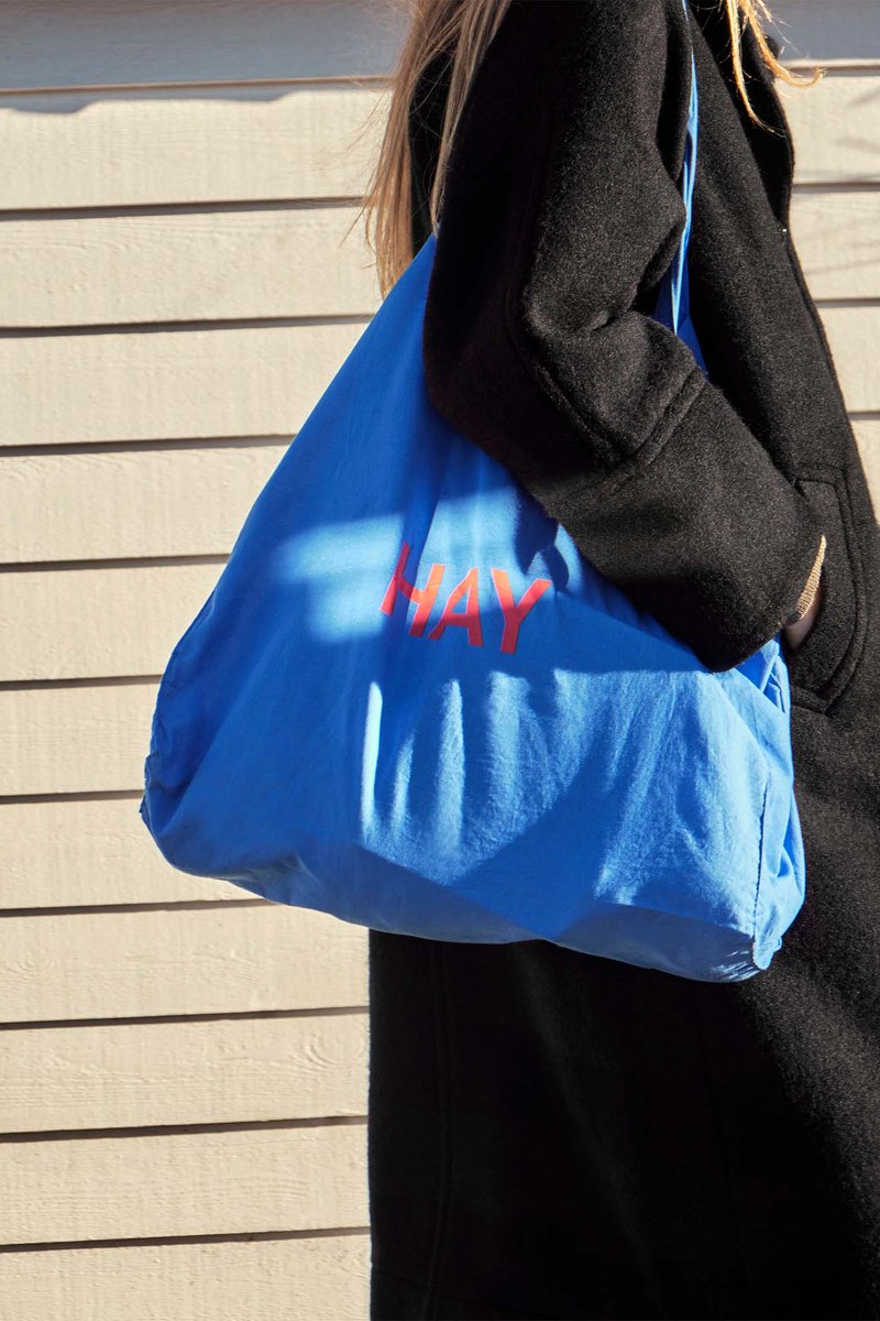 A person in a black coat carries the HAY Blue Tote Bag, Red Logo by HAY while walking past a beige wooden wall in sunlight.
