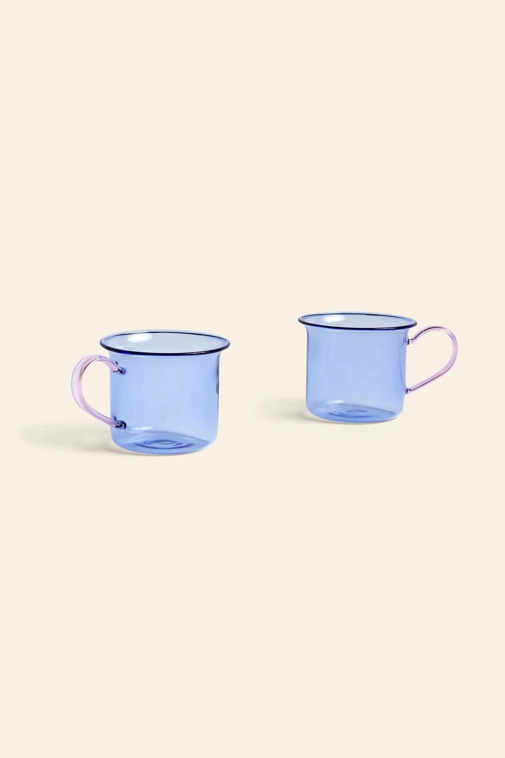 The HAY Borosilicate Cup, Set of 2 by HAY features transparent blue borosilicate glass with light pink handles and a modern design, adding a stylish touch when displayed side by side on a light beige background.