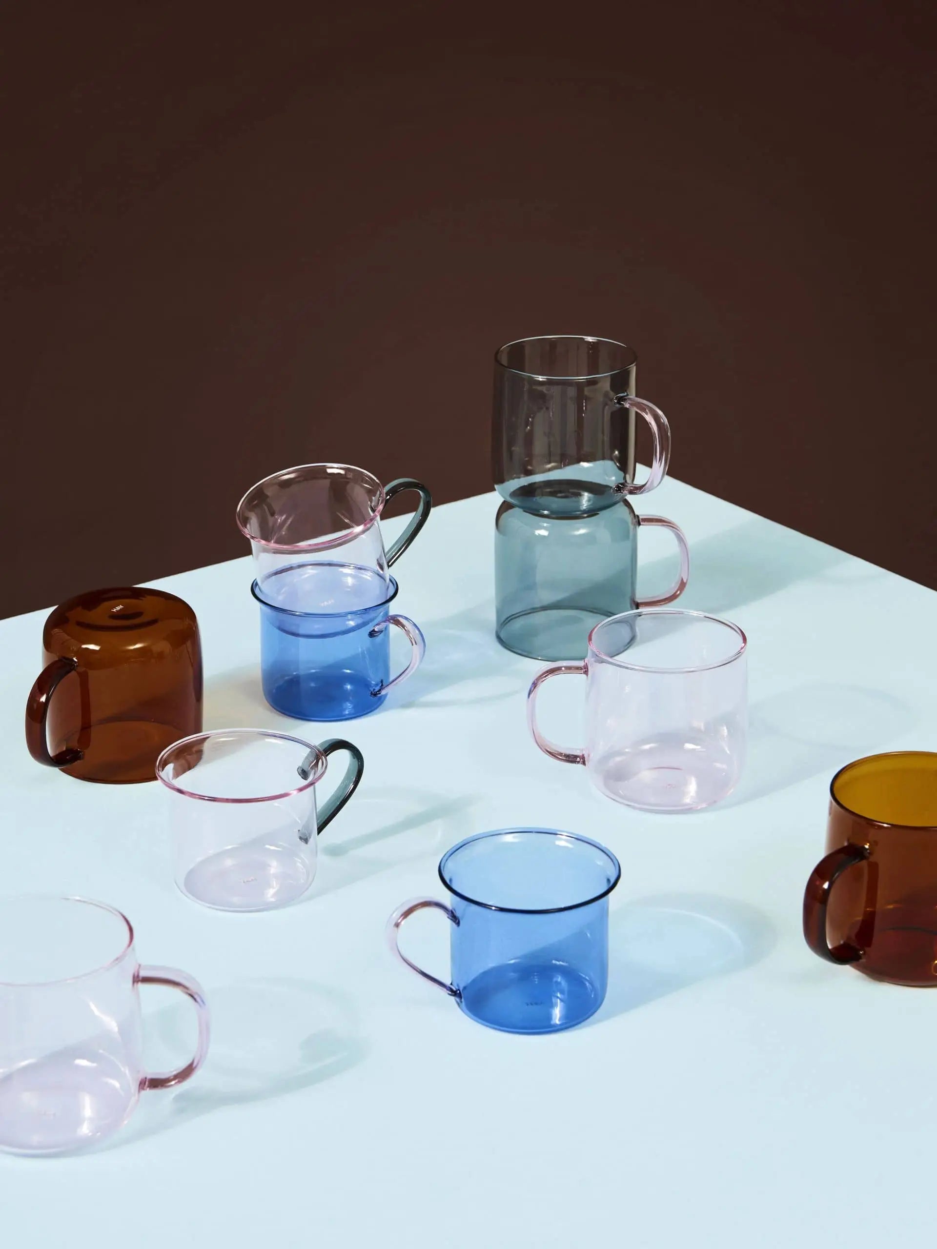 A set of HAY Borosilicate Cups in brown, blue, pink, and gray from HAY is displayed on a pale blue surface with a dark background; some cups stand upright while others lie on their sides.