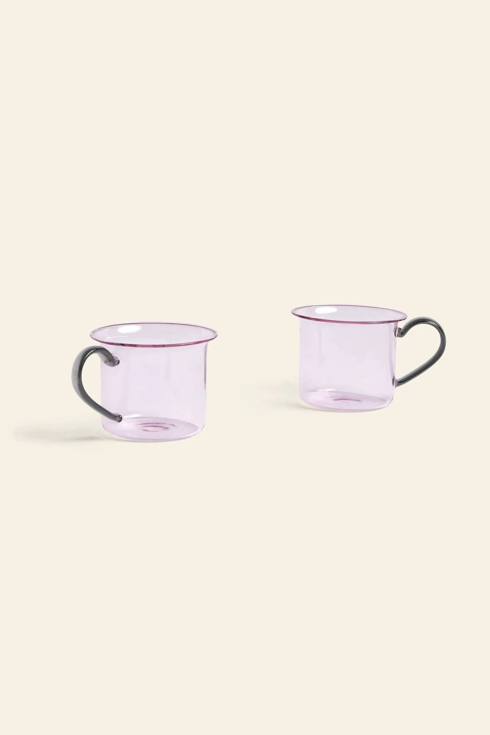 Two transparent light purple HAY Borosilicate Cups from HAY, each with a dark curved handle, are set apart on a light beige background. This is the HAY Borosilicate Cup, Set of 2.