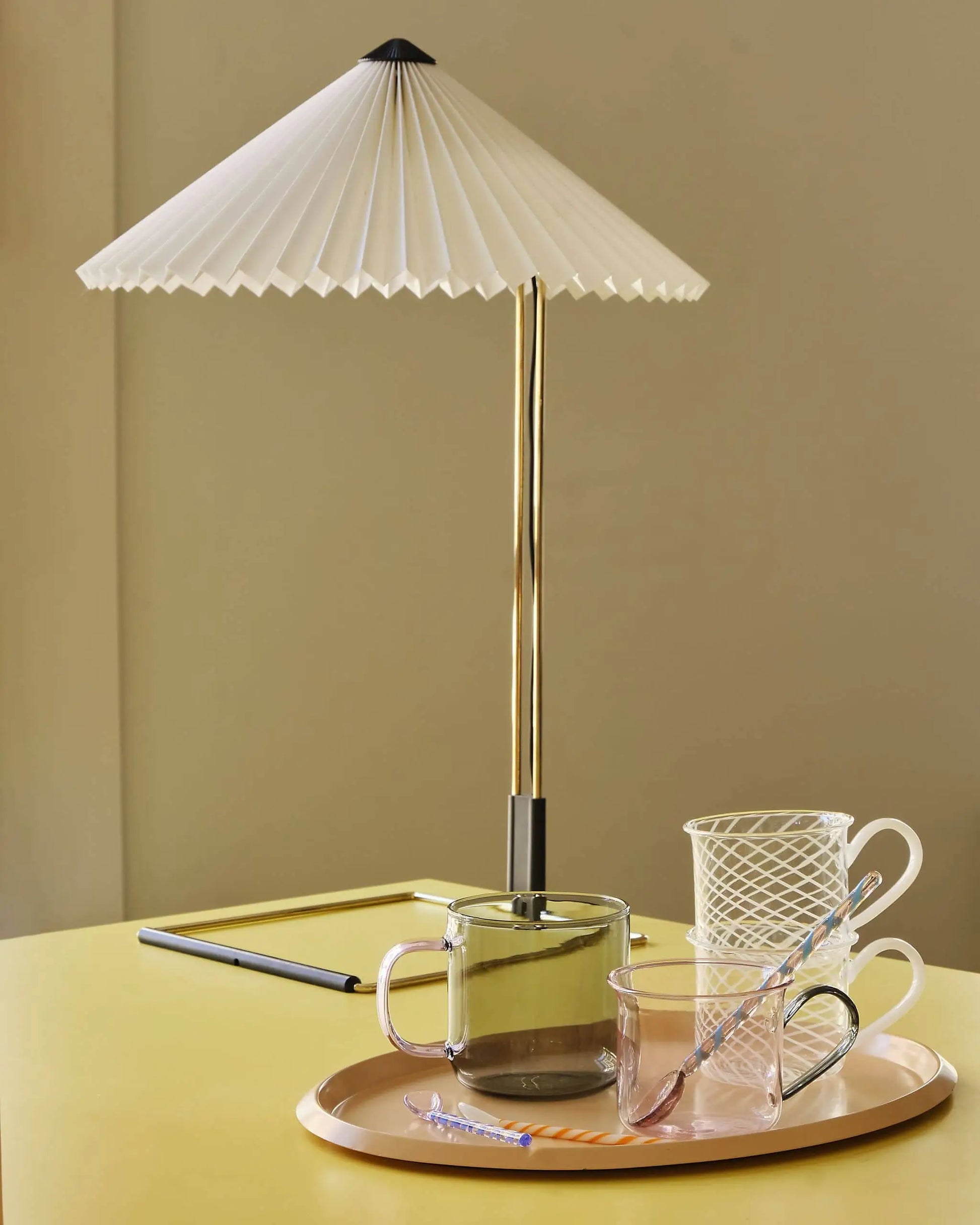 A modern table lamp with a pleated white shade stands on a yellow surface beside the HAY Borosilicate Cup, Set of 2 by HAY and two striped straws on a round tray, creating a minimalist and stylish arrangement.