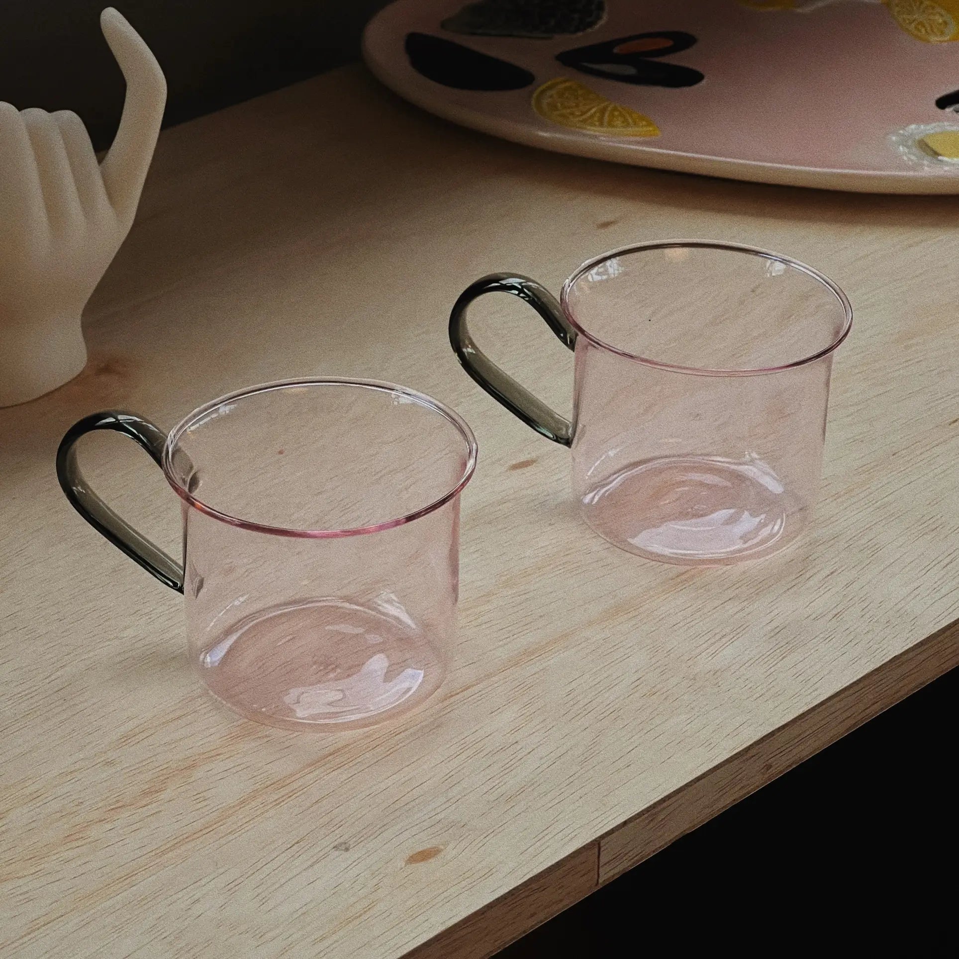 Two transparent pink HAY Borosilicate Cups (Set of 2) with dark handles by HAY rest on a light wood surface. Behind them are a fruit-themed decorative tray and a white hand-shaped object.