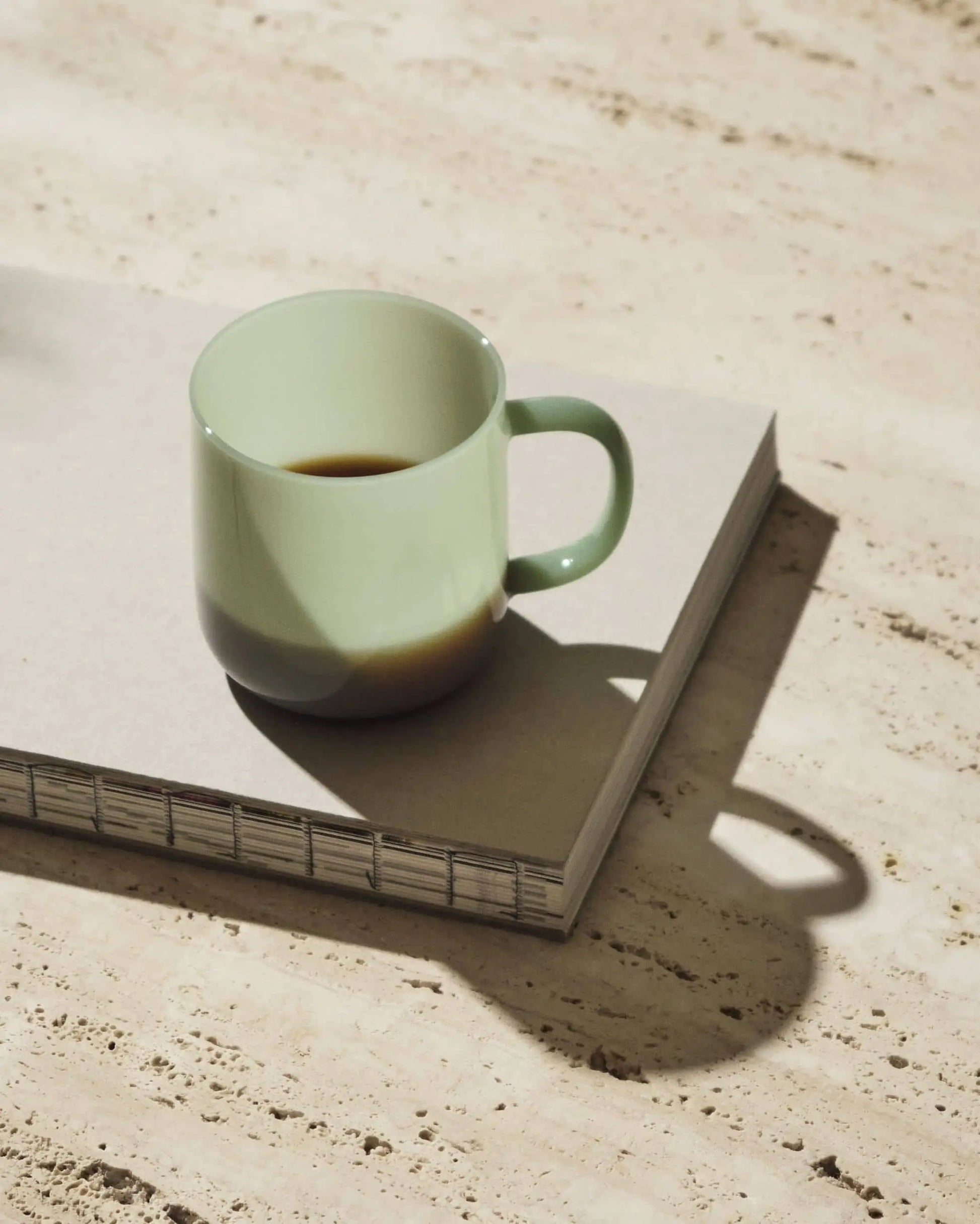 A lightweight HAY Borosilicate Mug from the Set of 2 by HAY in pale green, filled with dark coffee, rests on a closed spiral-bound notebook and casts a soft shadow on a light, textured surface.