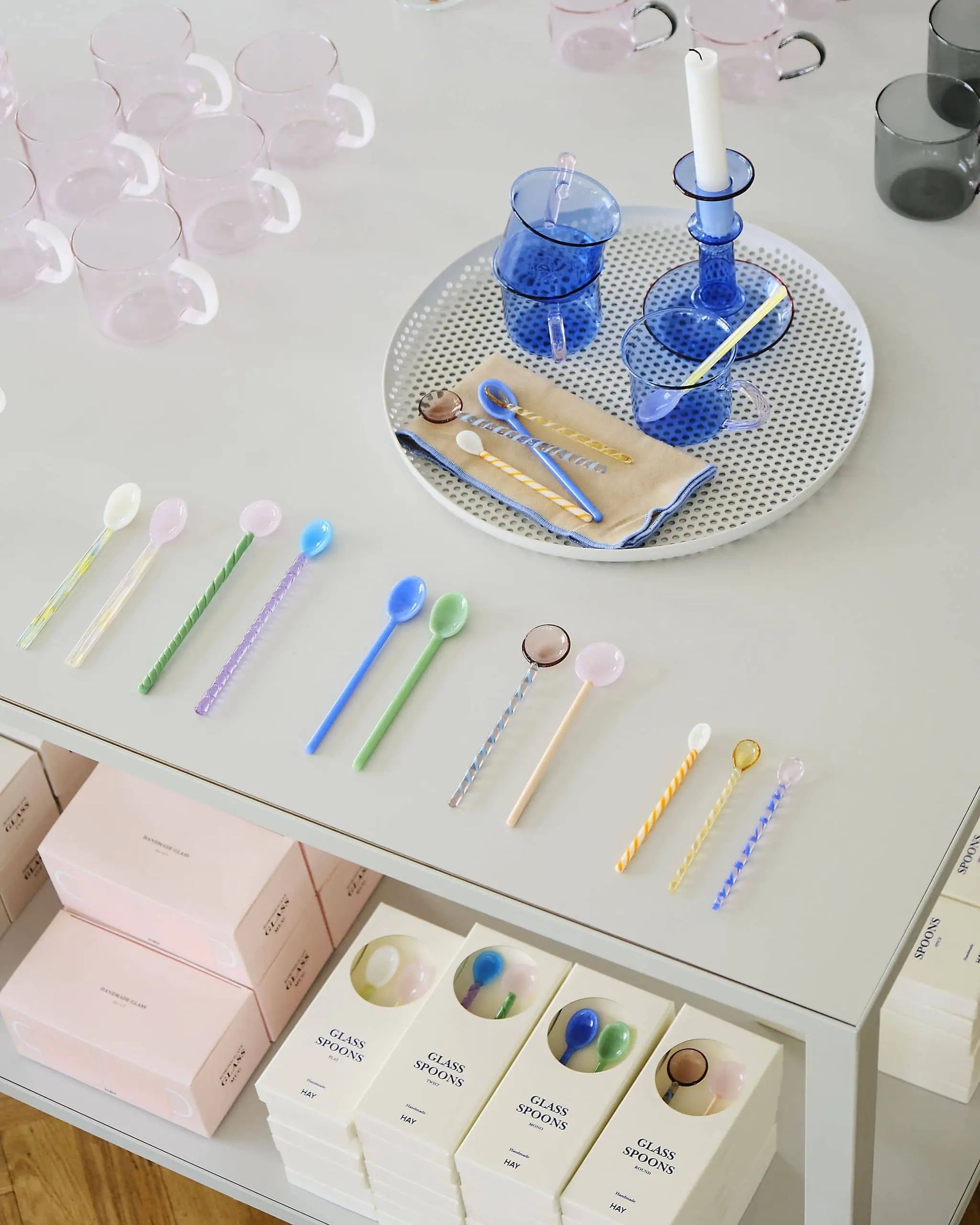 Colorful glass spoons are lined up on a white table, with boxes beneath them. Next to them are HAY Borosilicate Mug Sets of 2—lightweight, heat resistant mugs from HAY, ideal for daily use in pink and clear styles.