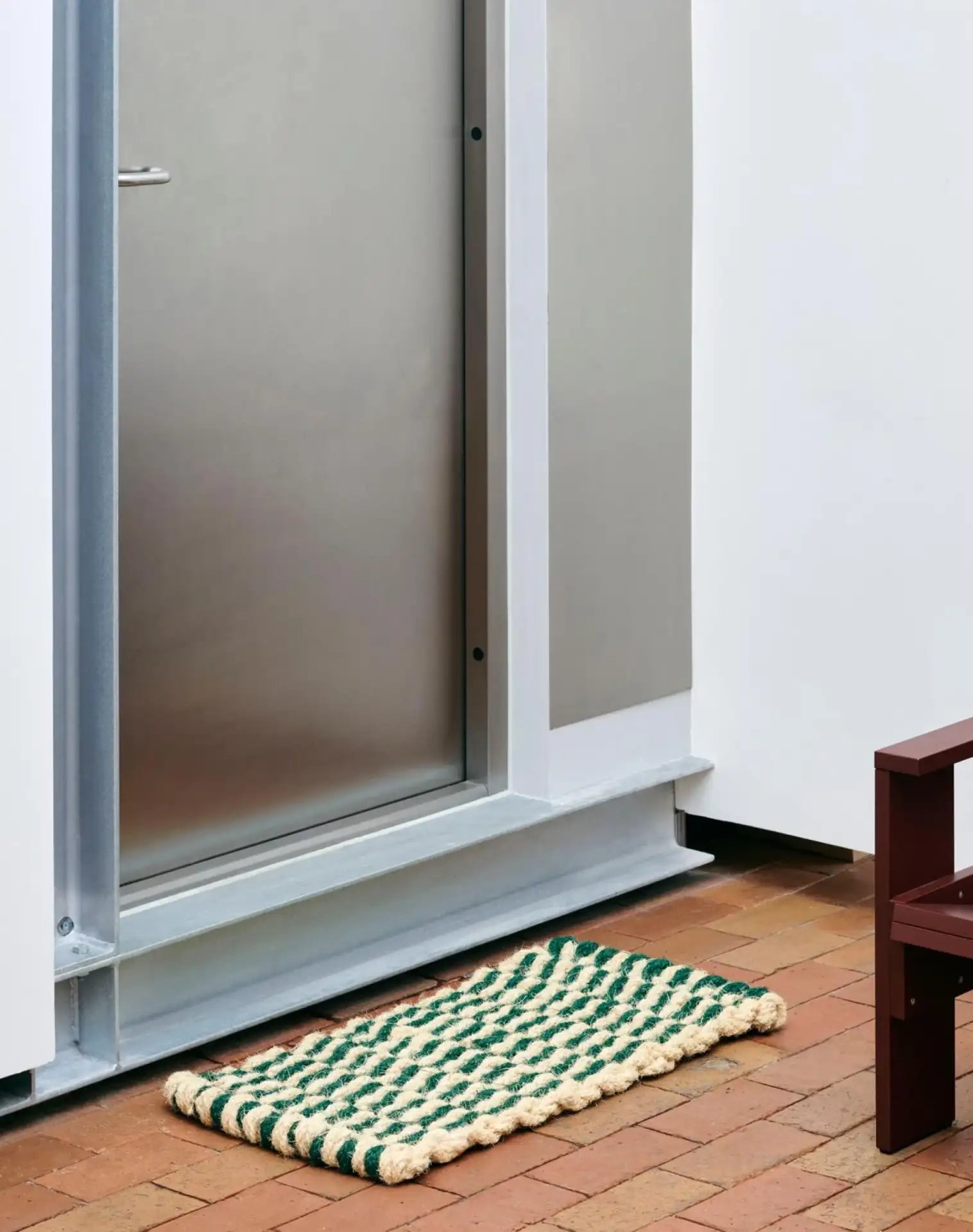 The HAY Coco Door Mat Stripe Wave by HAY, featuring green and cream stripes in durable coir, rests on a brick floor outside a metal door beside a white wall and brown chair.