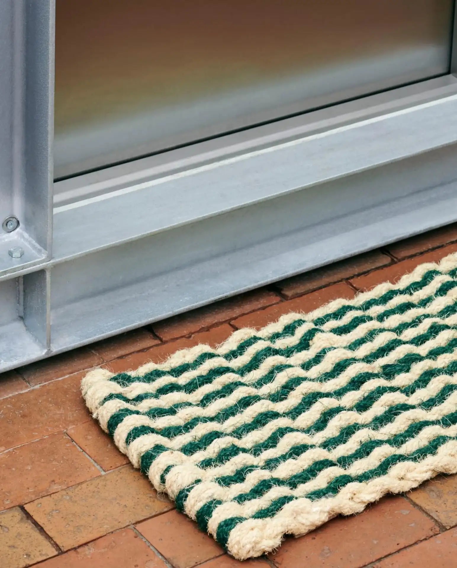 A close-up of the HAY Coco Door Mat Stripe Wave by HAY, featuring green and beige stripes in durable coir fibers, placed on a reddish-brown brick floor beside a metal door frame and glass door.