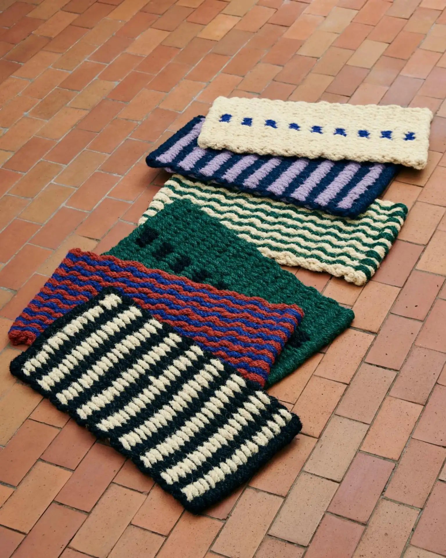 Five HAY Coco Door Mat Stripe Wave rugs by HAY, made with coir fibers, display colorful stripe and wave patterns. They are arranged diagonally on a reddish-brown tiled floor.