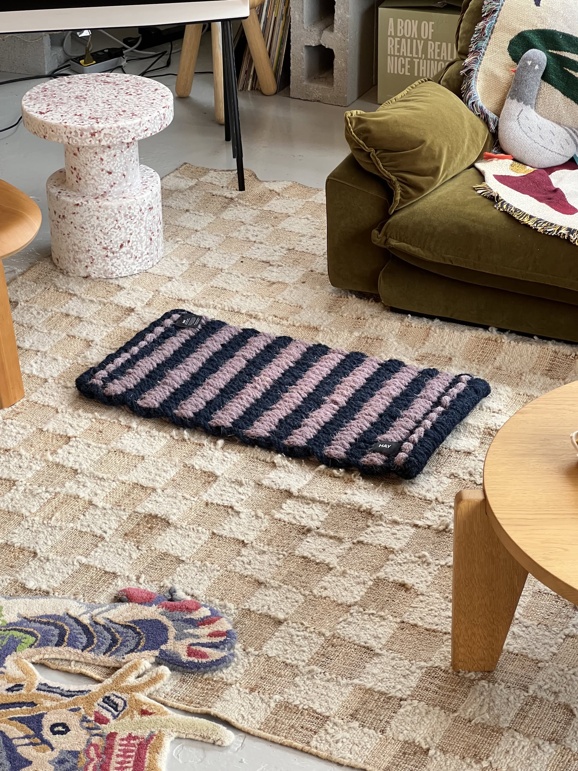 A cozy living room featuring a textured beige rug, the HAY Coco Door Mat Stripe Wide in navy and pink by HAY, a green sofa with a decorative pillow, a terrazzo side table, and a wooden coffee table on a patterned floor.