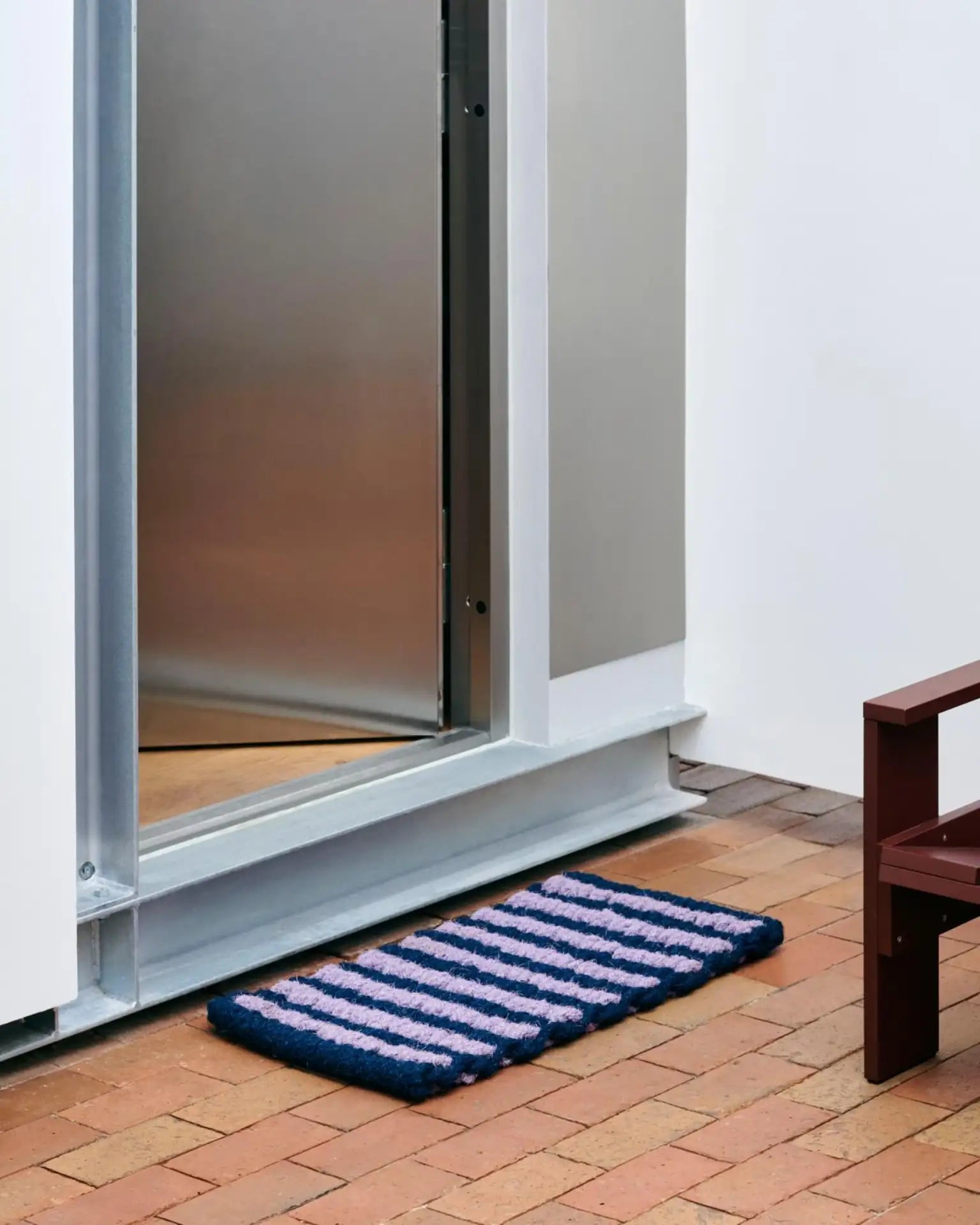 The HAY Coco Door Mat Stripe Wide by HAY, featuring blue and pink stripes and made from sturdy coir fibers, lies on a brick floor before a slightly open metal door with part of a brown bench visible on the right.