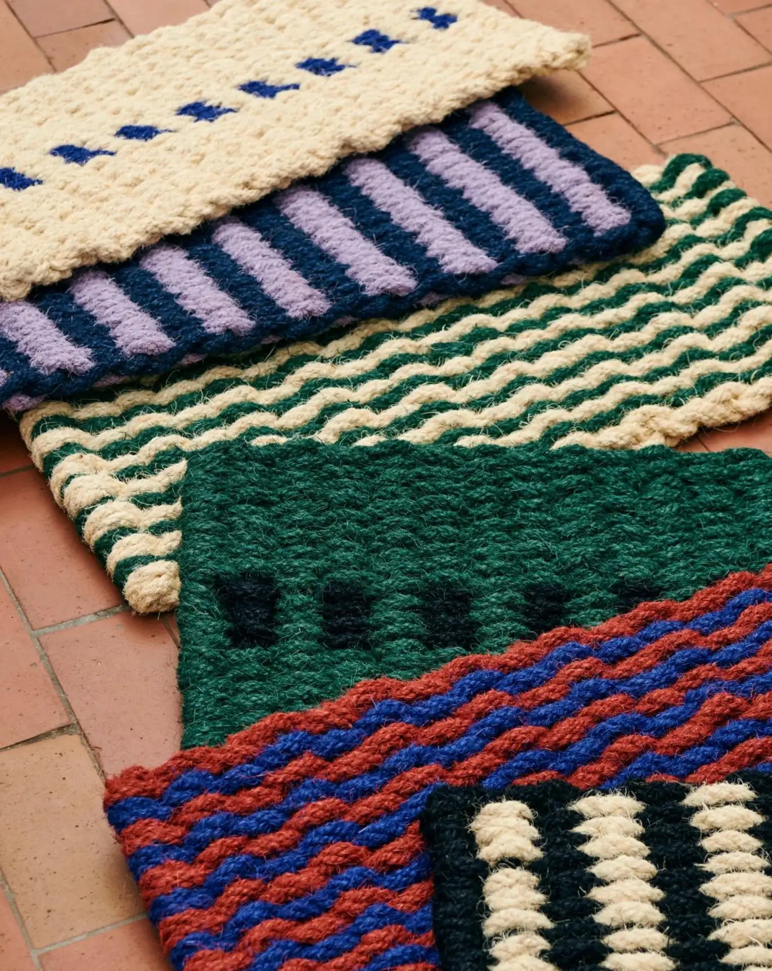 Four HAY Coco Door Mat Stripe Wide mats by HAY, featuring wavy and striped patterns in cream, blue, green, purple, and red, are arranged side by side on a terracotta tiled floor.