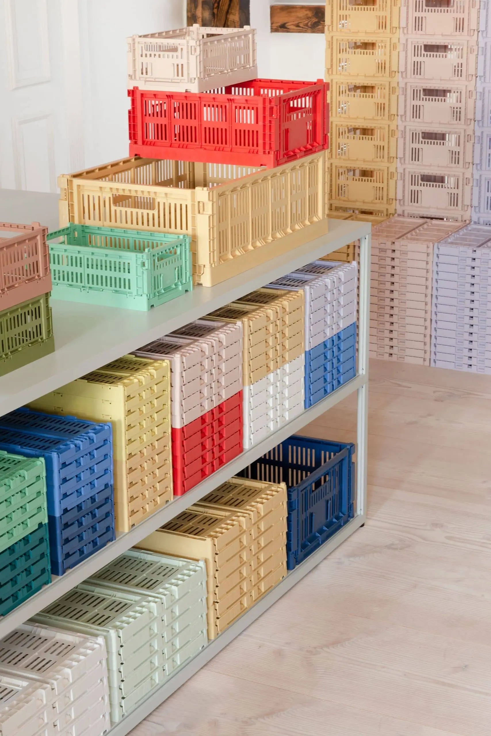 Brightly stacked HAY Colour Crate Small storage boxes by HAY in red, yellow, green, blue, and beige sit on metal shelves and tables atop light wood flooring, creating an organized and colorful living space.