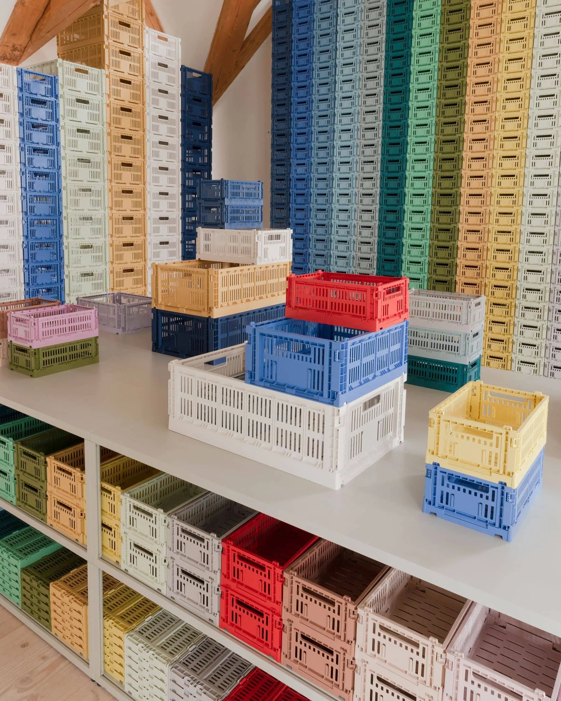 A display of HAY Colour Crate Small stackable plastic boxes by HAY arranged on shelves and a table showcases pastel and bright colors, creating a vibrant, organized living space with tall stacks reaching the ceiling.
