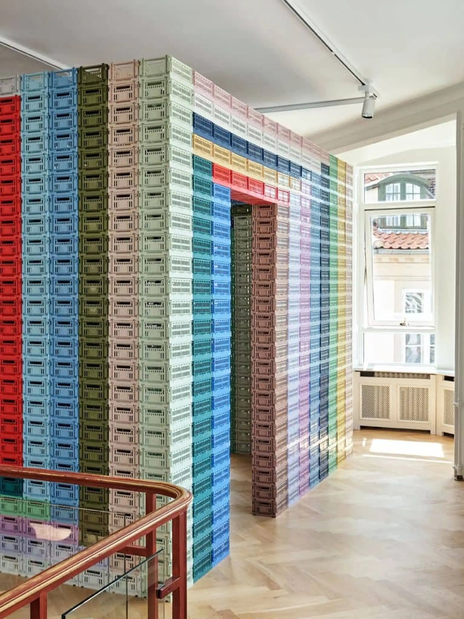 A room with walls made of colorful, stacked HAY Colour Crate Small storage units by HAY, arranged in playful patterns around a doorway and large windows, letting natural light brighten the organized wooden-floored space.