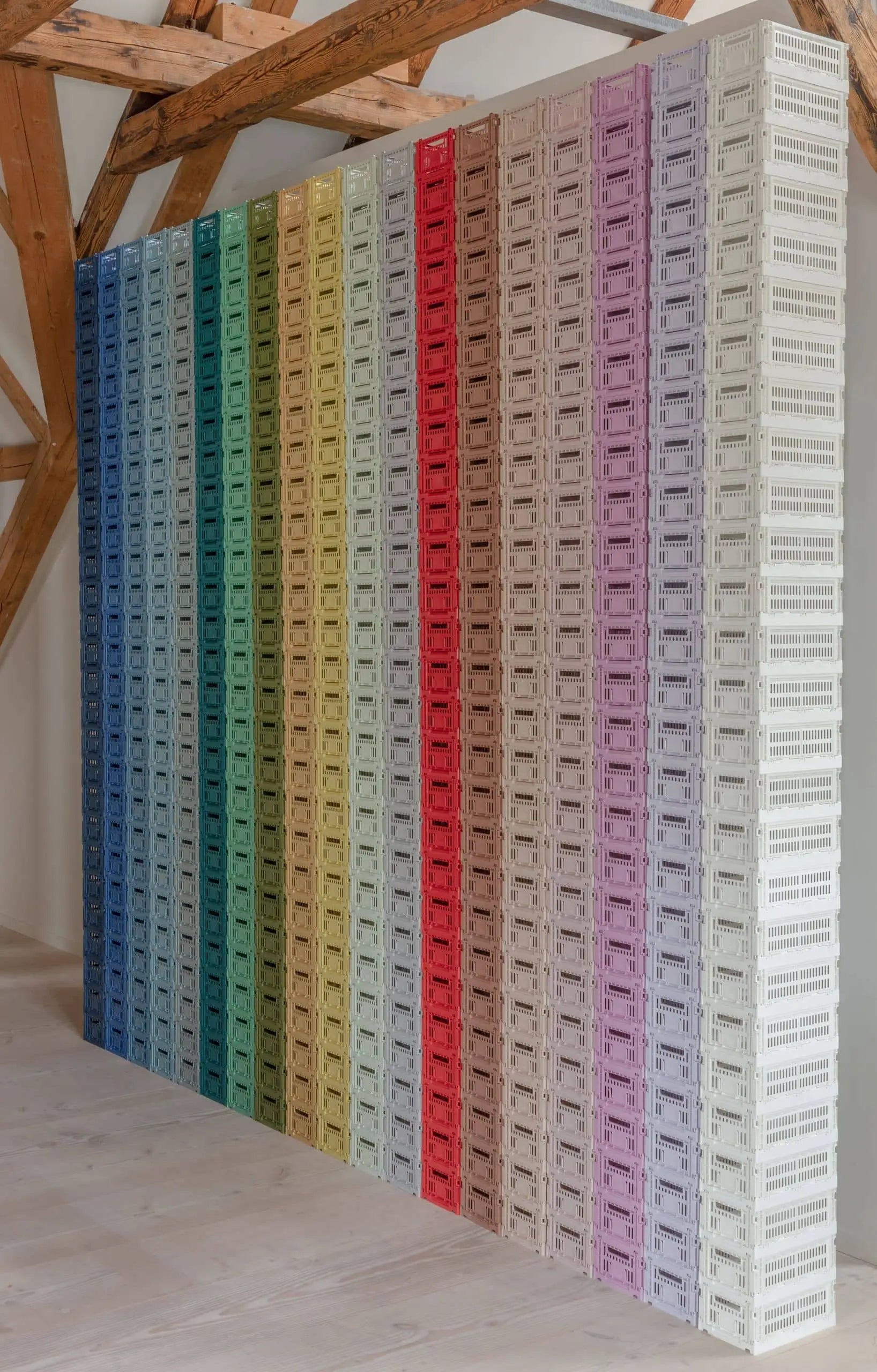 A wall of HAY Colour Crate Small by HAY, stacked in vertical rows to create a blue-to-white gradient, adds vibrant storage to a room with wooden beams and light flooring.