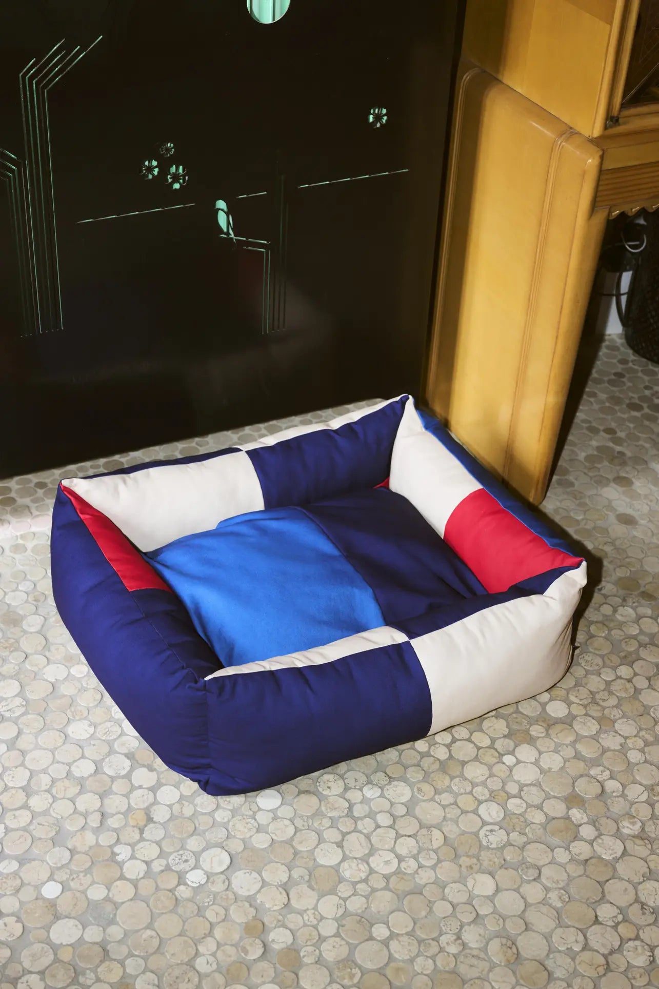 The HAY Dogs Bed M by HAY—a square, padded bed made from recycled polyester in blue, white, and red—rests on a pebble-patterned tile floor near a yellow chair and black surface, with a blue blanket inside.