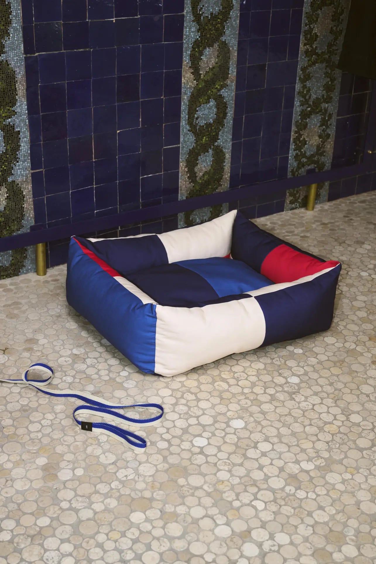 A square HAY Dogs Bed M by HAY, made from recycled polyester in red, blue, white, and black sits on a pebble-tiled floor near a blue tiled wall. A blue leash is placed next to the bed.