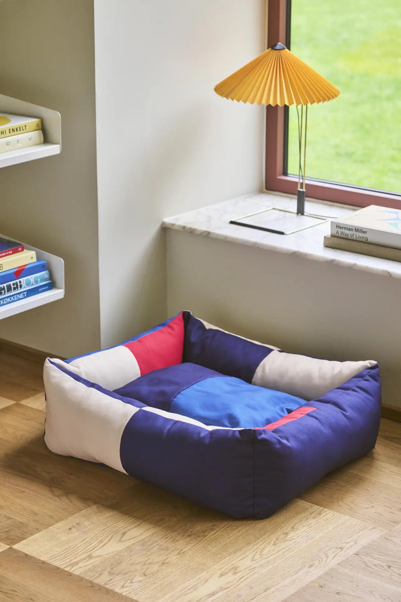 The HAY Dogs Bed M, a colorful square bed by HAY made from recycled polyester, sits on a wooden floor near a window with a marble sill, beside a yellow lamp and a bookshelf filled with books.