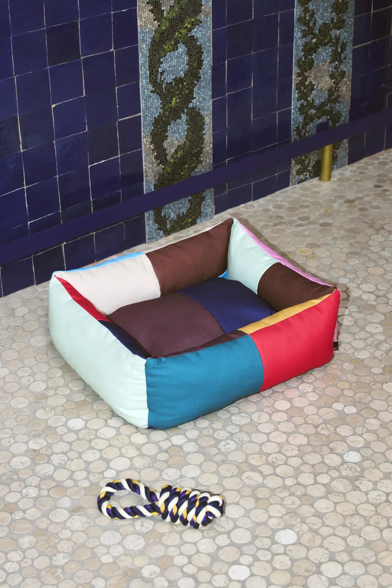 The HAY Dogs Bed S by HAY, crafted from recycled polyester in a colorful patchwork design, sits on a pebble-patterned floor with a twisted rope dog toy. Blue tiled walls with snake-like mosaic add to the lively backdrop.