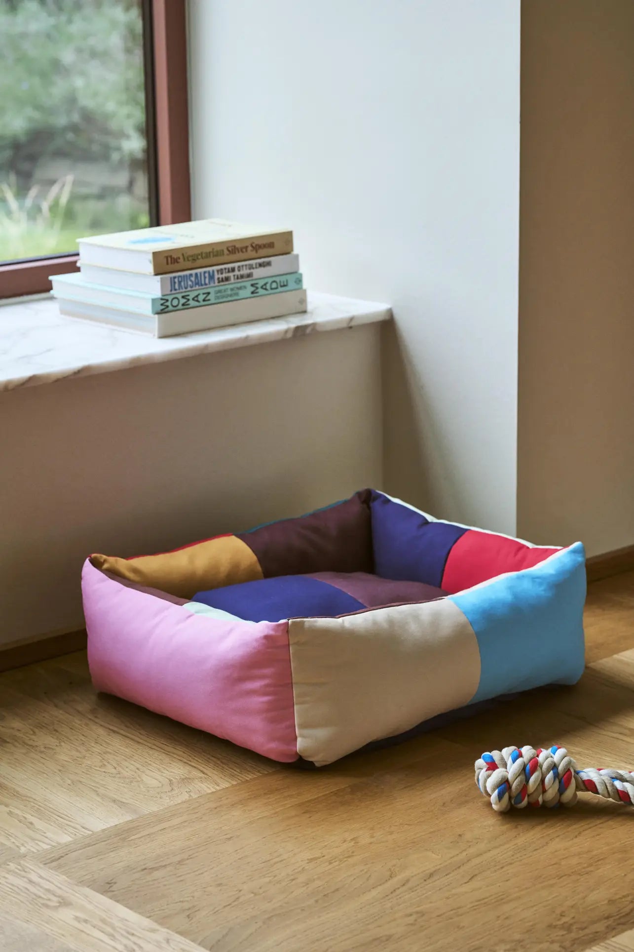 The HAY Dogs Bed S by HAY, crafted from recycled polyester in a colorful patchwork design, rests on a wooden floor near a window, accompanied by stacked books on the sill and a multicolored rope toy nearby.