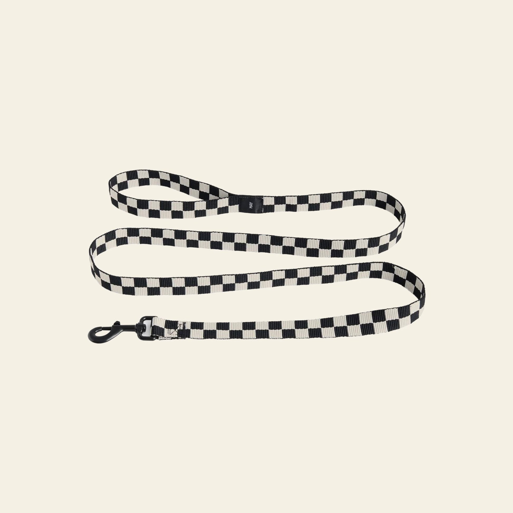 The HAY Dogs Leash Flat Check by HAY, made from recycled polyester in a black and white checkered pattern with a metal clasp, is shown in loose loops on a light beige background.