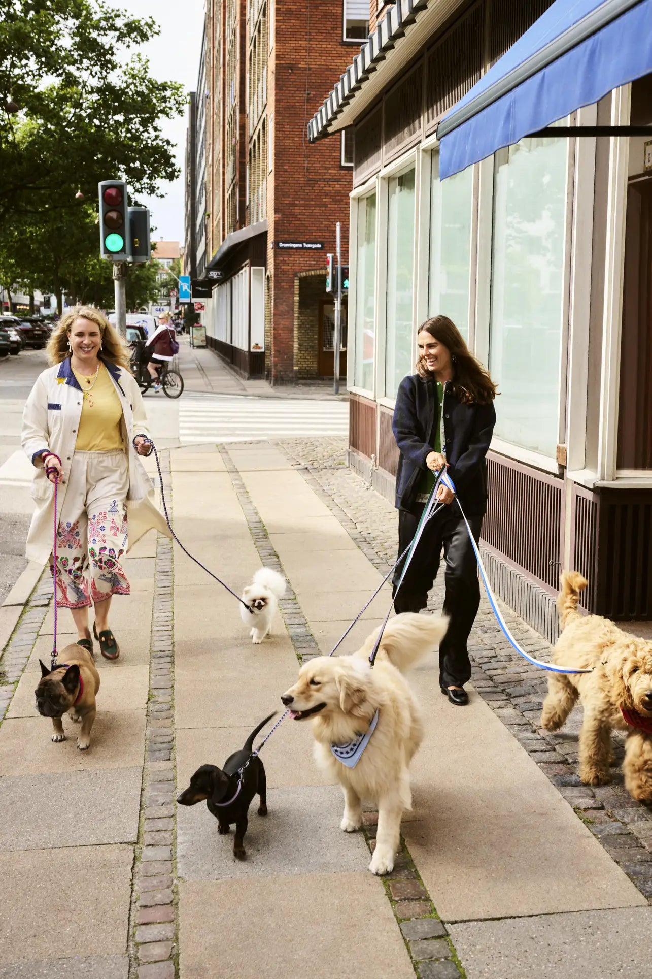Two women walk several dogs on a city sidewalk, one holding the HAY Dogs Leash Flat M/L by HAY made from recycled polyester, as buildings line the street and a cyclist rides by in the background.