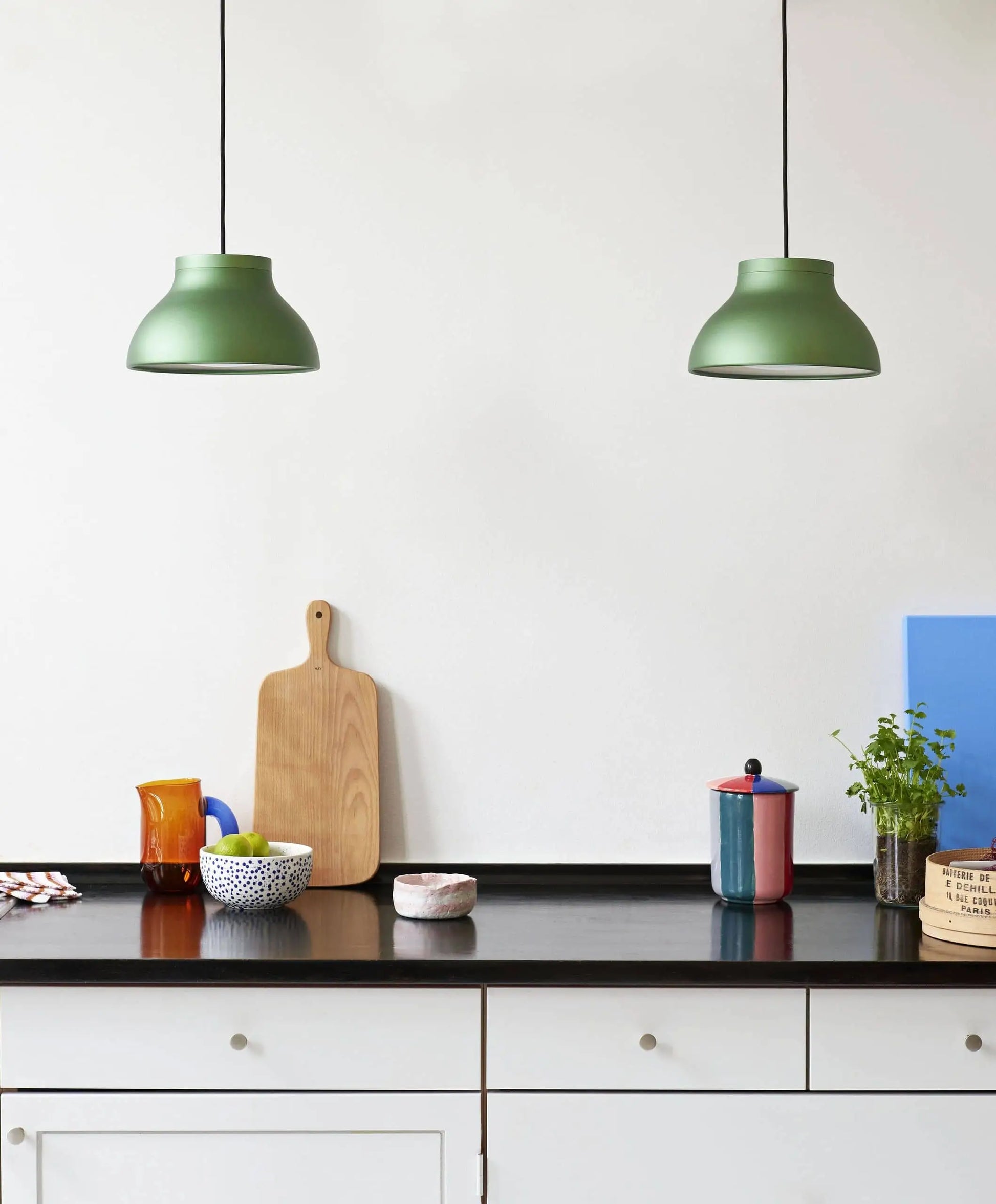 Modern kitchen counter with a black surface, assorted dishes and jars, a colourful HAY Jug S by HAY for serving drinks, wooden and blue cutting boards, small potted plant, two green pendant lights above, and white cabinetry below.