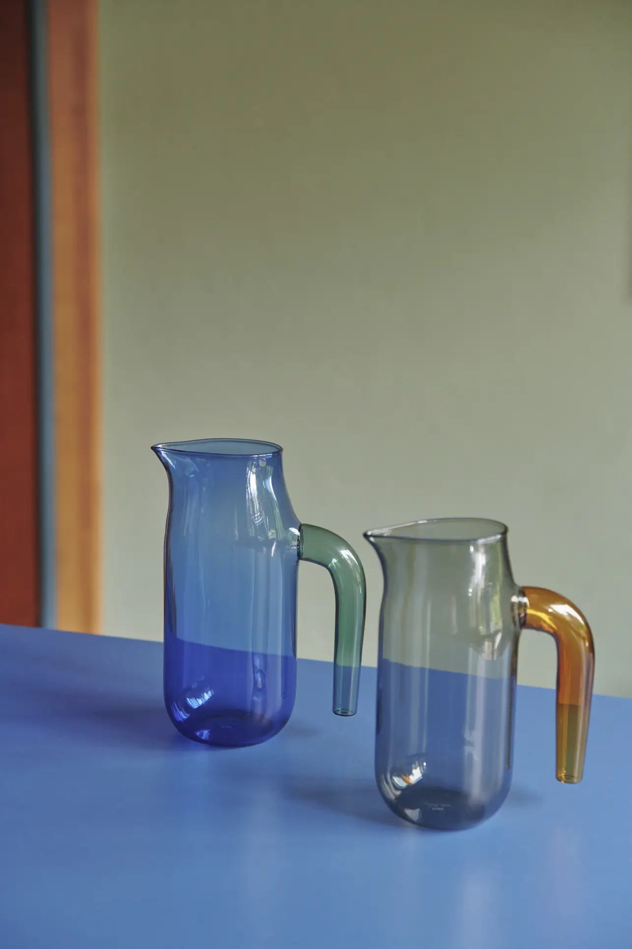 Two HAY Jug XL glass jugs by HAY sit on a blue surface—one blue with a green handle, the other clear gray with an amber handle—both crafted from borosilicate glass. The softly lit background shows muted green and tan hues.