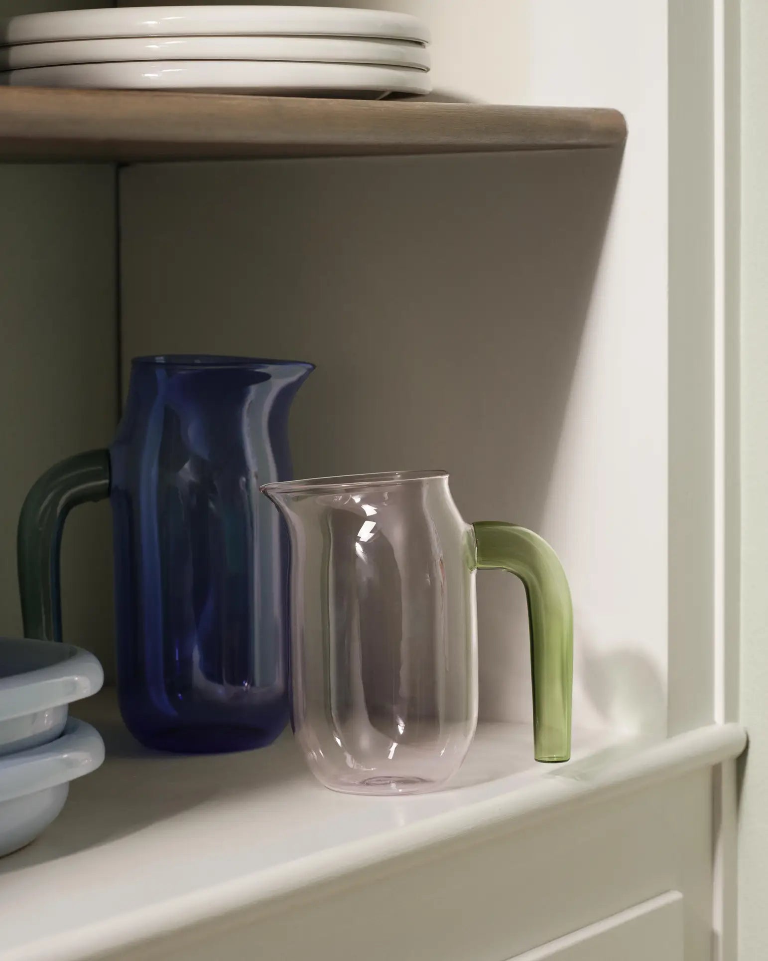 Two HAY Jug XL pieces by HAY—one blue with matching handle, the other clear with a green handle—made from borosilicate glass, sit on a white shelf below stacked plates and beside light blue containers.