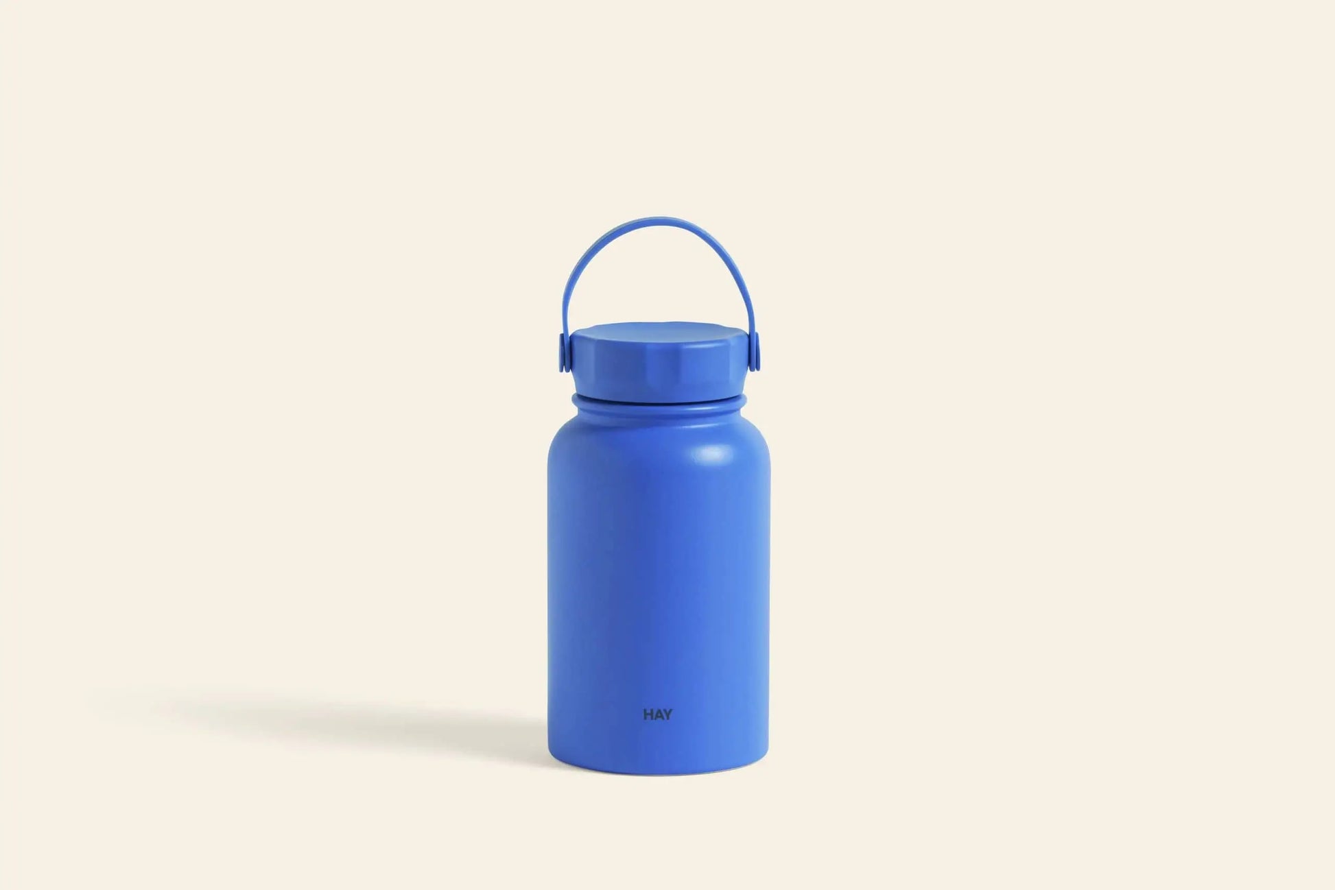 The HAY Mono Thermal Bottle by HAY, in stylish bright blue with a wide round body, screw-on lid, and matching handle, stands upright on a light beige backgroundโideal for keeping beverages fresh.