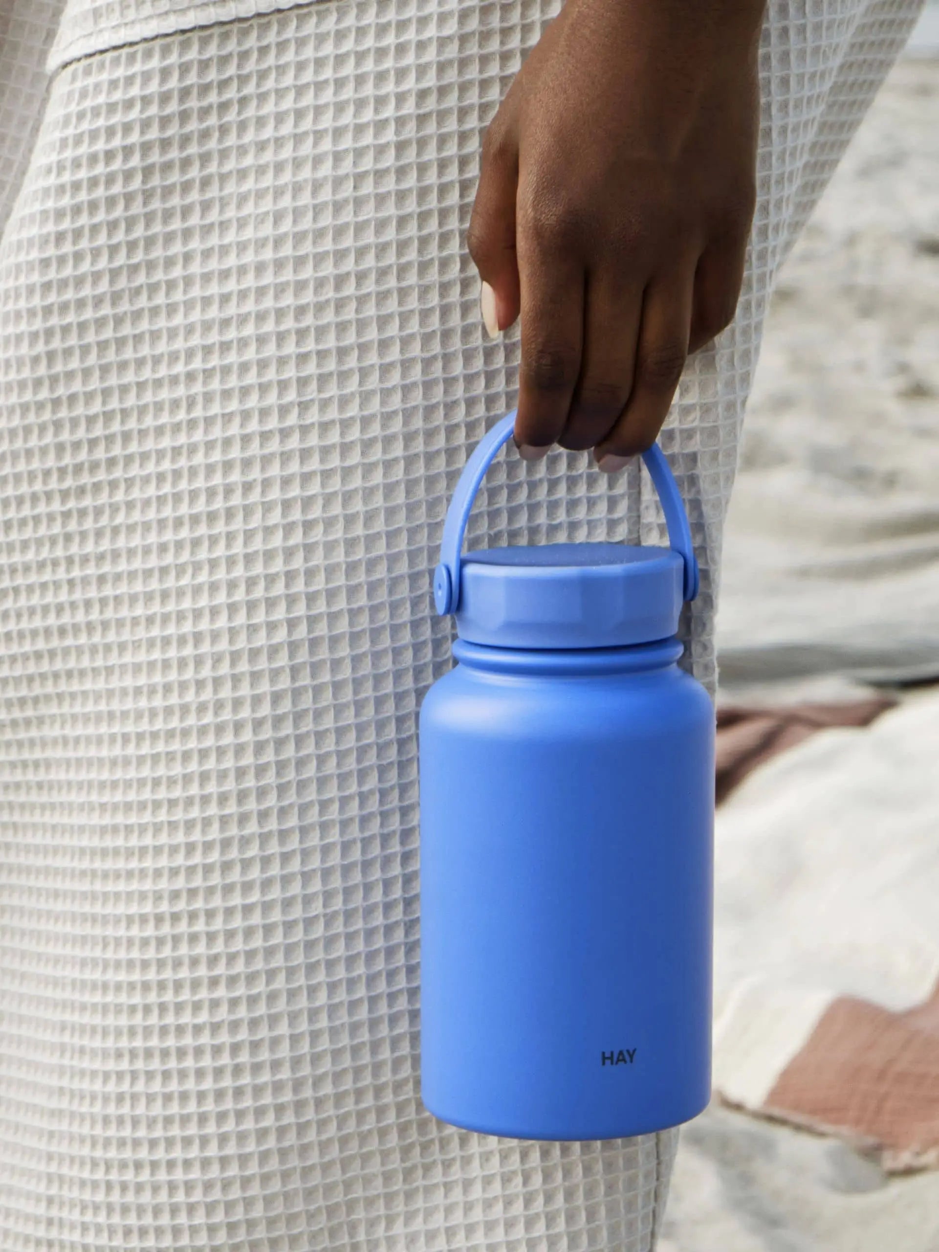 A stylish individual in a textured, light outfit holds the HAY Mono Thermal Bottle in matte blue with a handle, perfect for keeping drinks cool. The bottle features HAY near the bottom, set against sandy and striped fabric backgrounds.