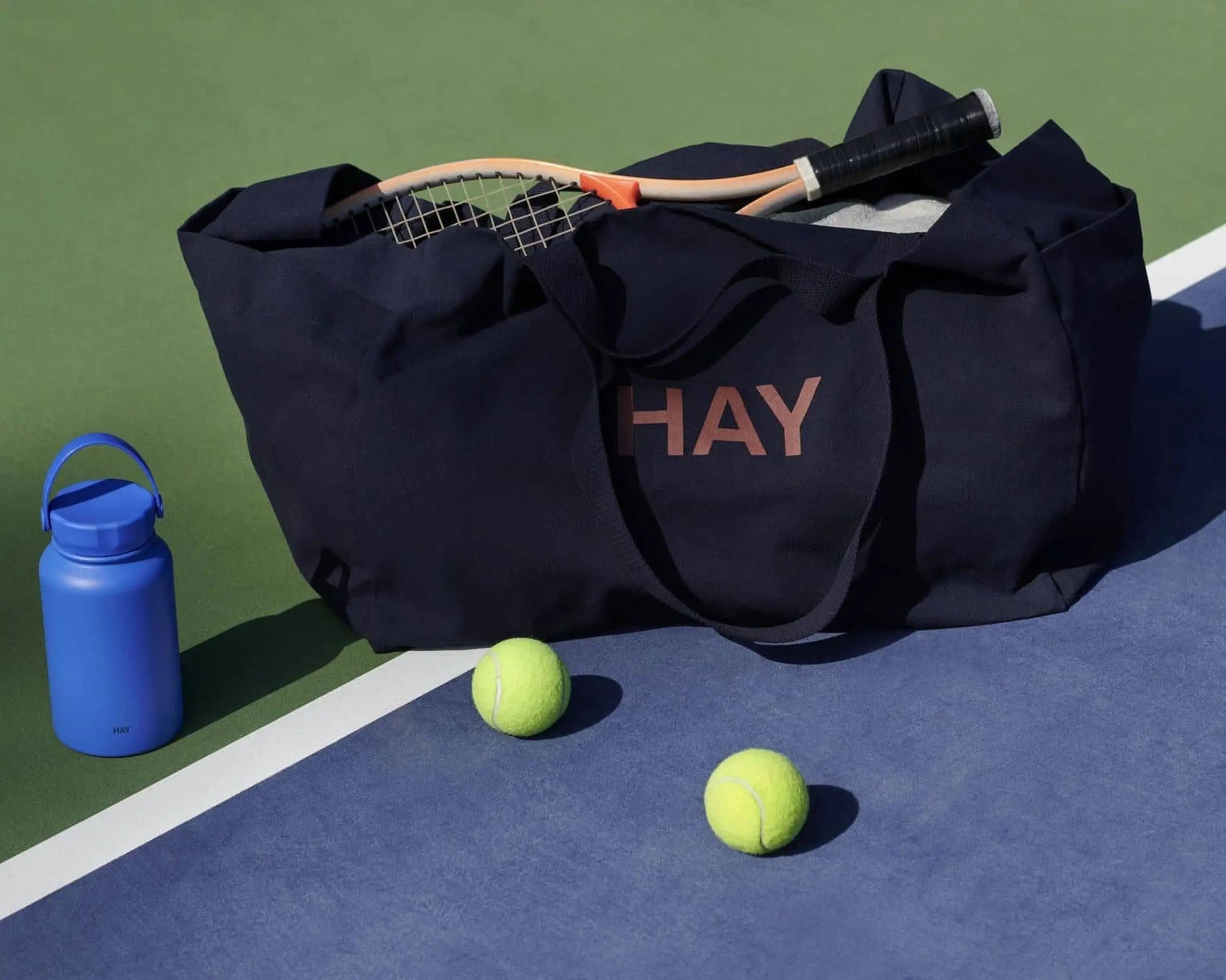 A navy blue HAY sports bag with a tennis racket rests on the court beside a blue HAY Mono Thermal Bottle and two tennis balls.