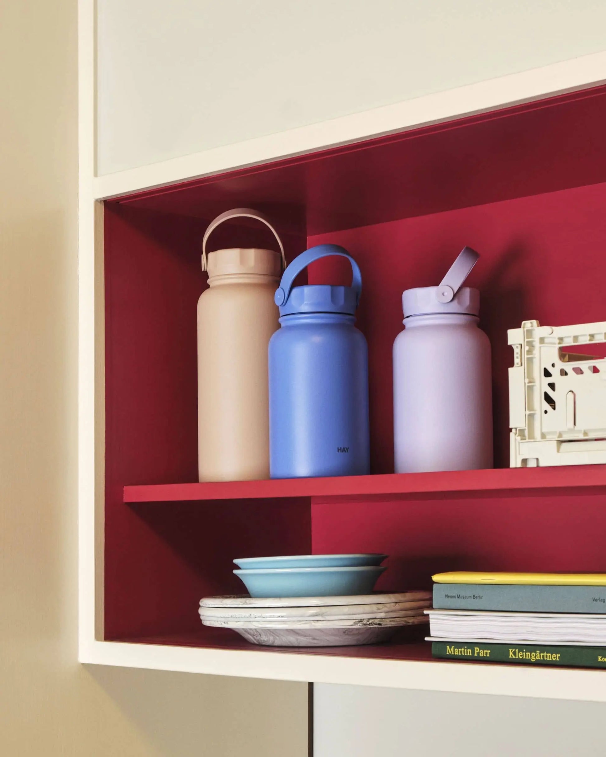 Three pastel HAY Mono Thermal Bottles in peach, blue, and lavender by HAY stand on a red shelf above plates and books in a white-framed cabinet, ready to keep your beverages at the ideal temperature.