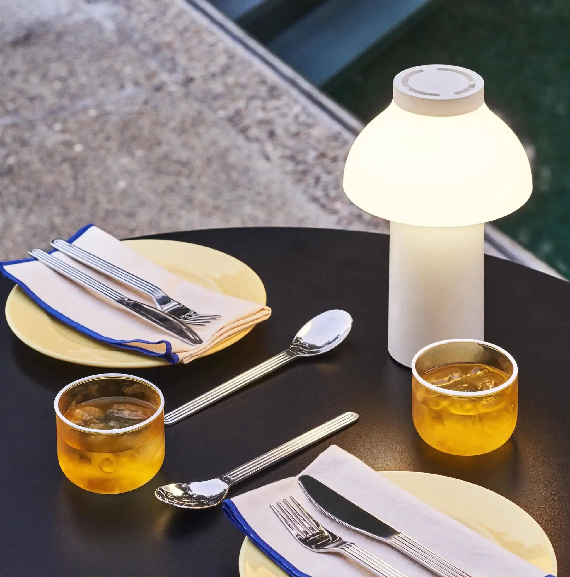 A round black table with two yellow plates, cutlery-wrapped napkins, iced drinks, and a HAY PC Portable Lamp by HAY sits near a concrete surface with water in the background.