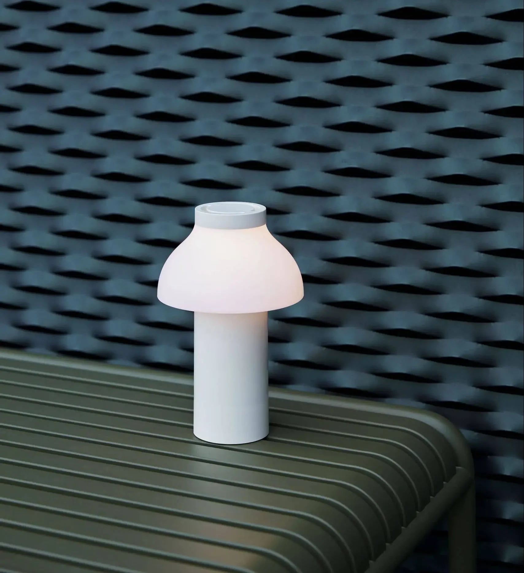 A compact, white HAY PC Portable Lamp by HAY glows softly on a ribbed olive-green bench, set against a textured dark blue wall.