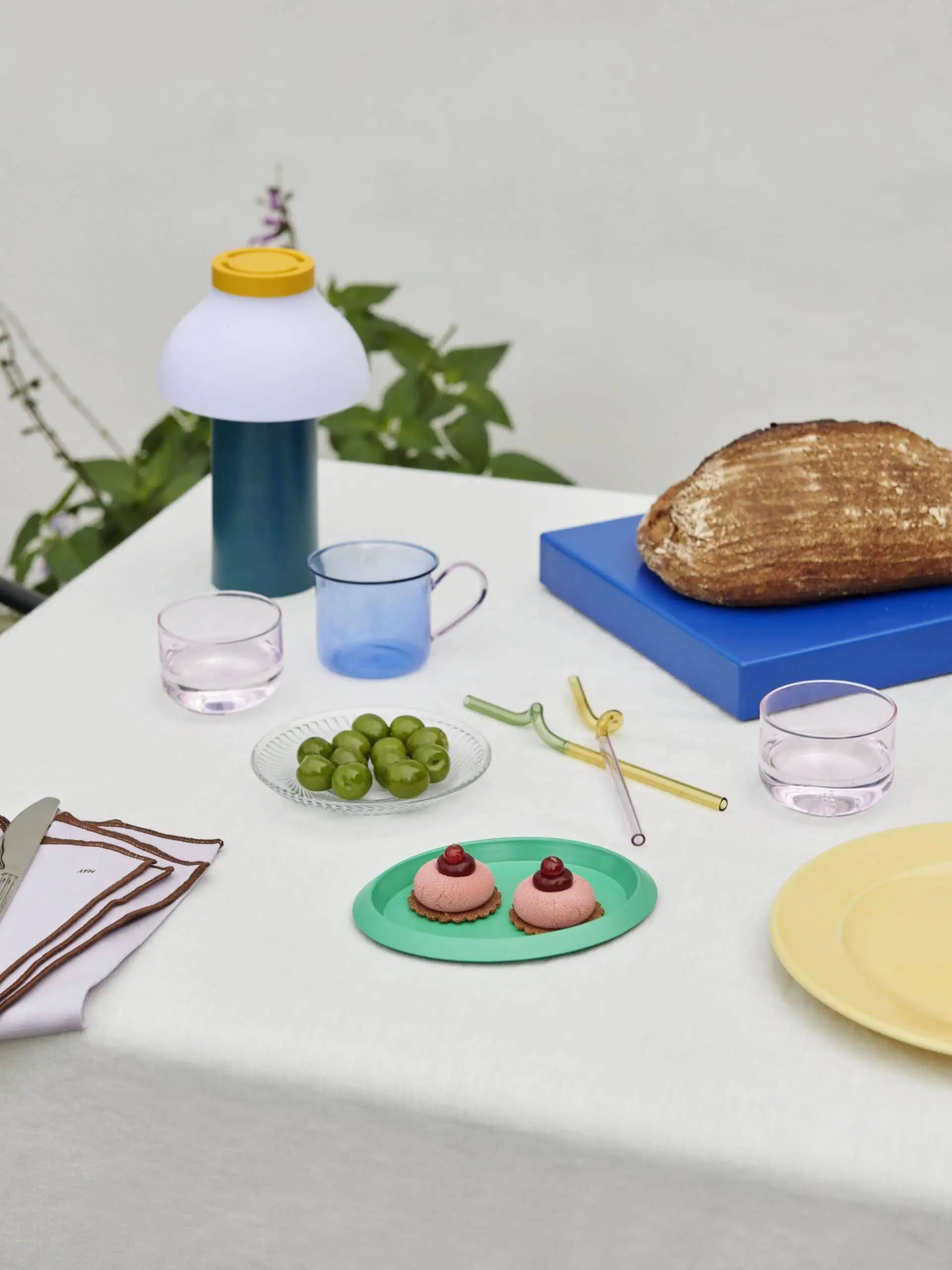 A table set with a loaf of bread on a blue block, two pink pastries on a green plate, pastel glasses, and the HAY PC Portable Lamp by HAY for an Art Deco touch, plus plates, straws, green grapes, utensils, and a yellow-capped bottle.