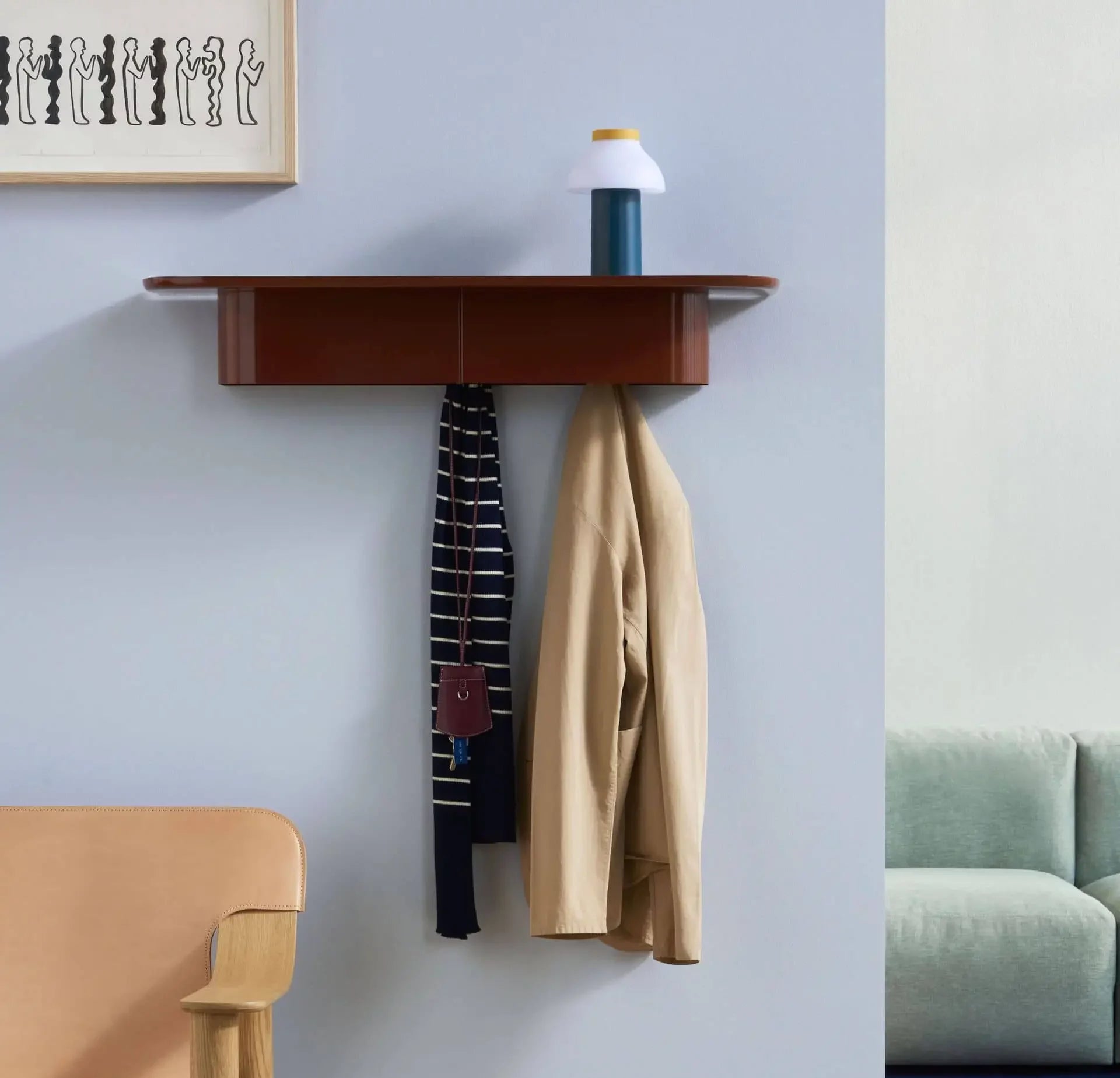 A wooden wall shelf holds a white and blue vase; below, a beige jacket, striped scarf with keys hang. Nearby are an abstract art piece, a HAY PC Portable Lamp by HAY, a tan chair, and part of a green sofa.