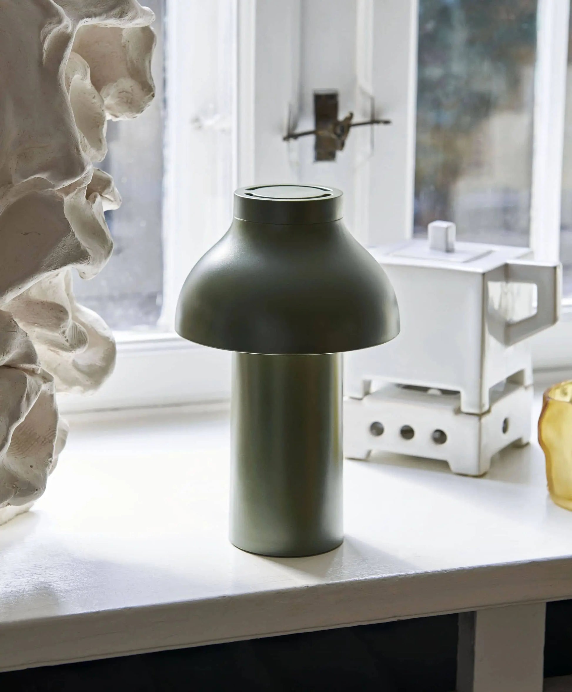 A matte green HAY PC Portable Lamp by HAY, featuring a cylindrical base and dome-shaped shade, sits on a white windowsill with abstract sculptures and geometric decor, combining subtle Art Deco touches with modern design.