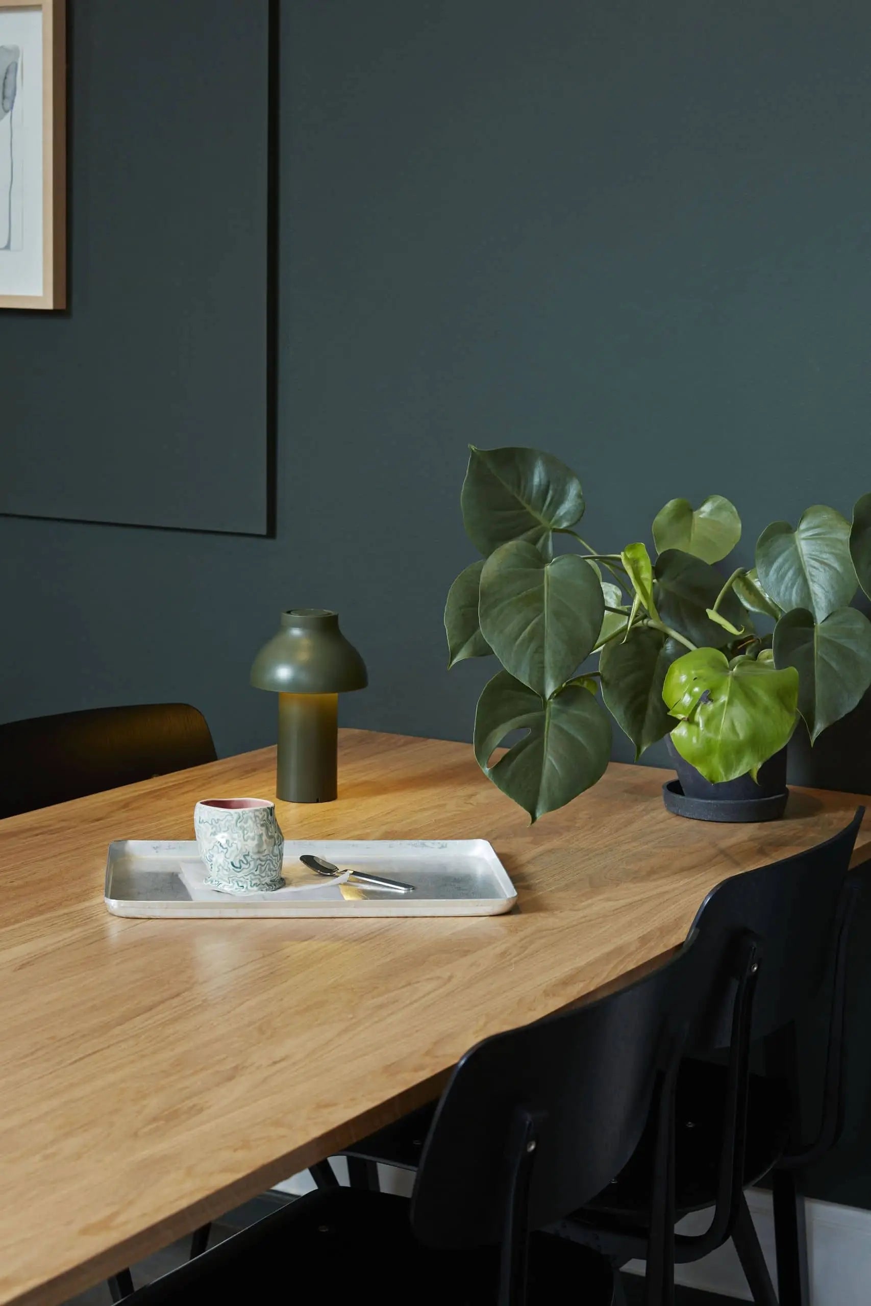 A modern wooden table with black chairs, a green potted plant, ceramic cup and plate on a tray, a green vase, and a HAY PC Portable Lamp by HAY in Art Deco style is set against a dark green wall.