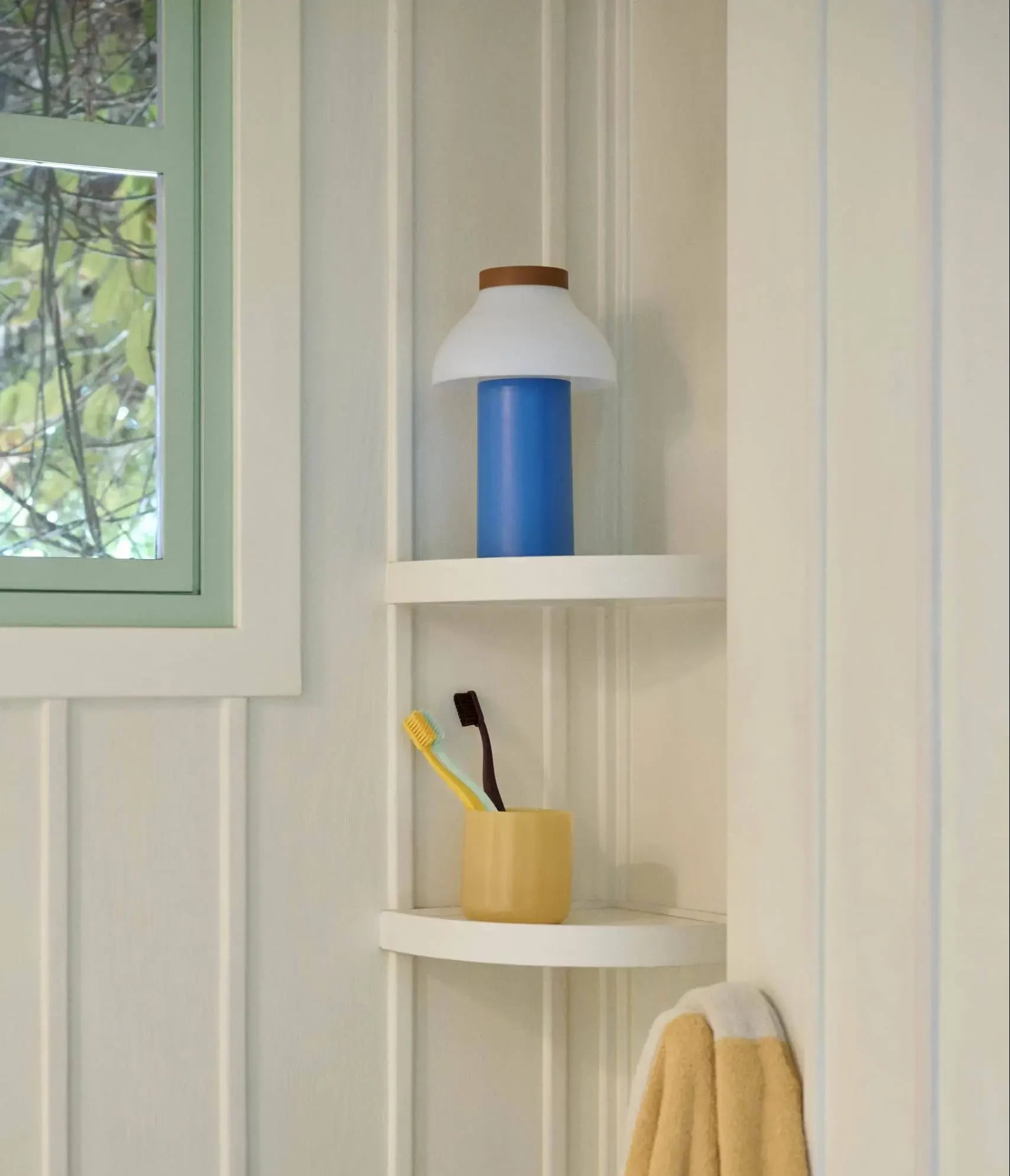 A bathroom corner shelf holds a HAY PC Portable Lamp in blue and white on the top shelf, with a yellow cup containing two toothbrushes below, near a yellow towel on a hook. A green-framed window is visible in the background.