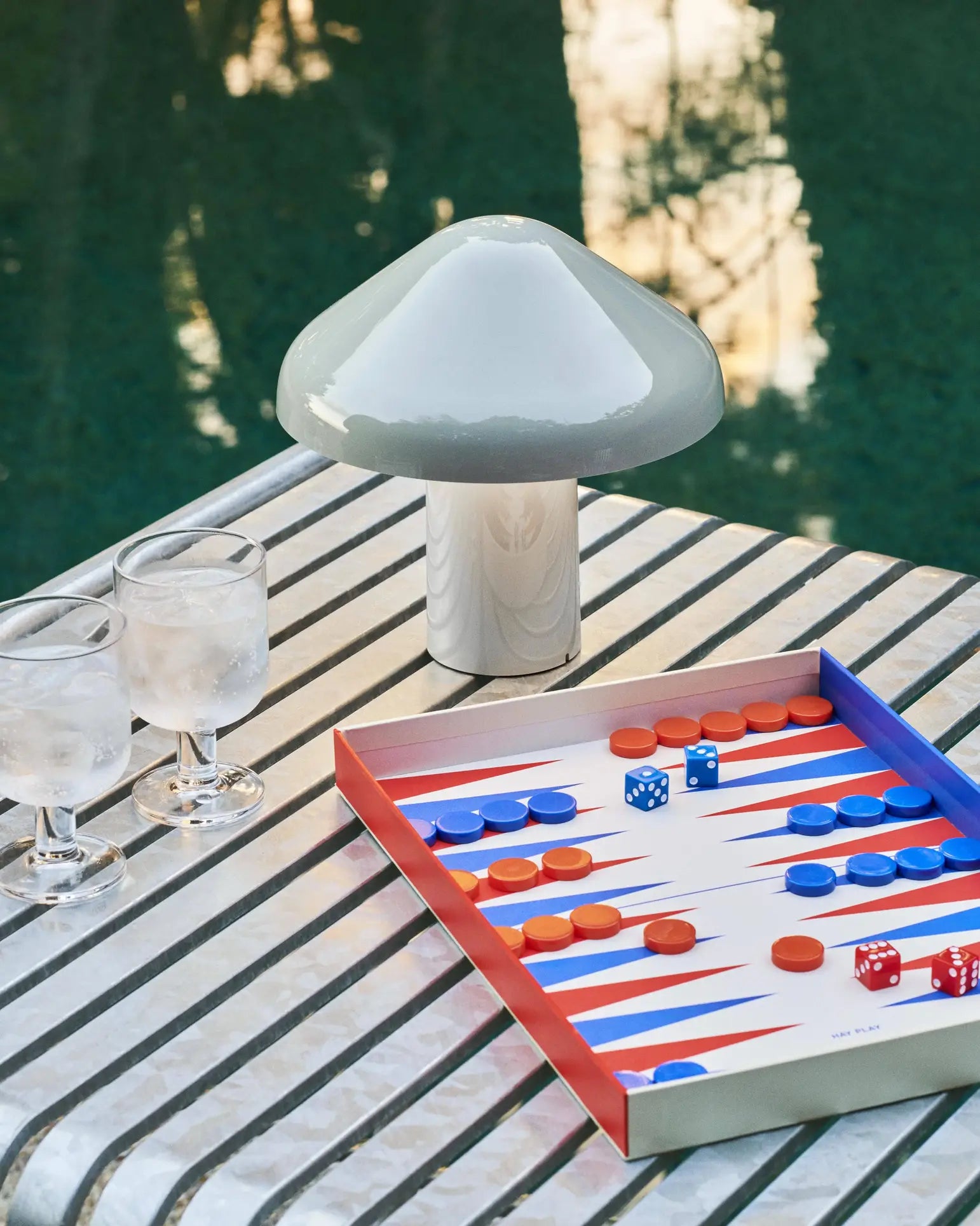 A striped poolside table holds two water glasses, an open backgammon set with red and blue pieces and dice, and a white HAY Pao Portable Lamp by HAY.