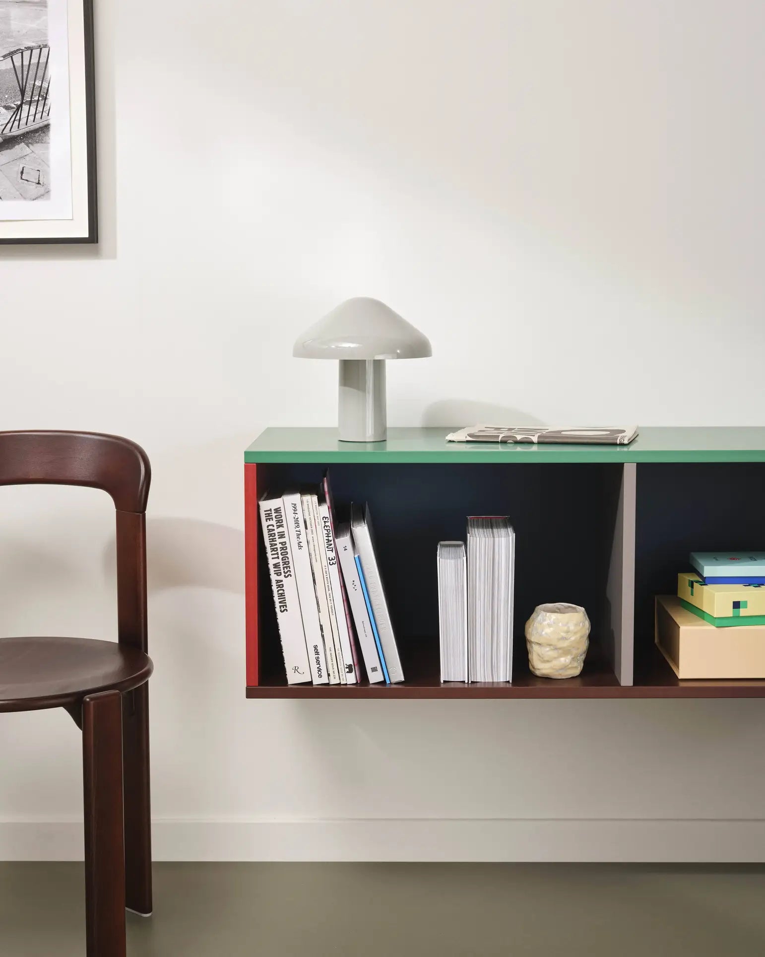 A modern minimalist room features a green floating shelf with books, magazines, boxes, a decorative object, and the HAY Pao Portable Lamp by HAY. A wooden chair sits nearby, and part of a framed artwork is visible on the wall.