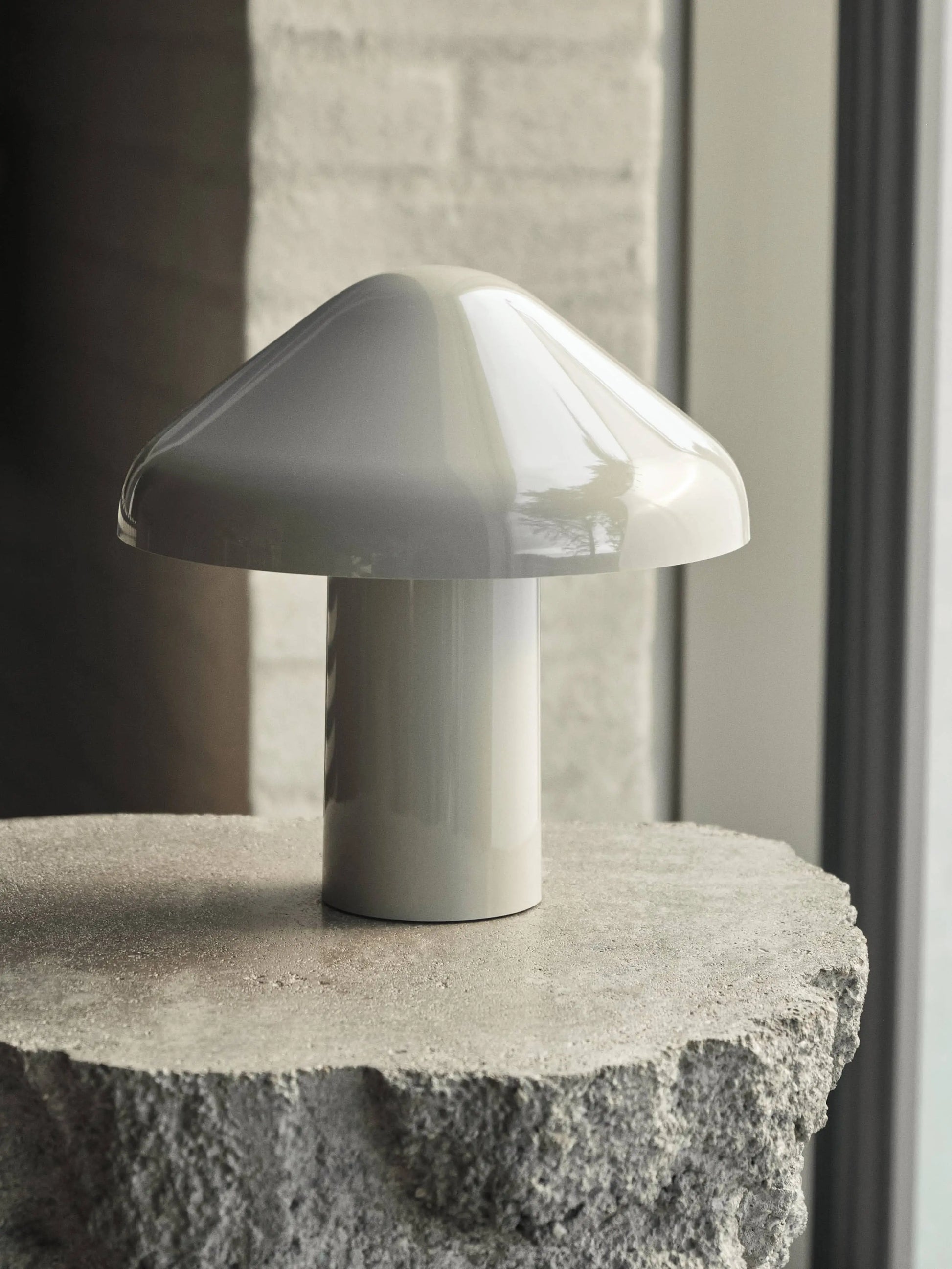 The HAY Pao Portable Lamp by HAY, with its glossy white mushroom-shaped shade and base, sits on a rough stone surface near a window, showcasing its sleek, modern portable design in natural light.