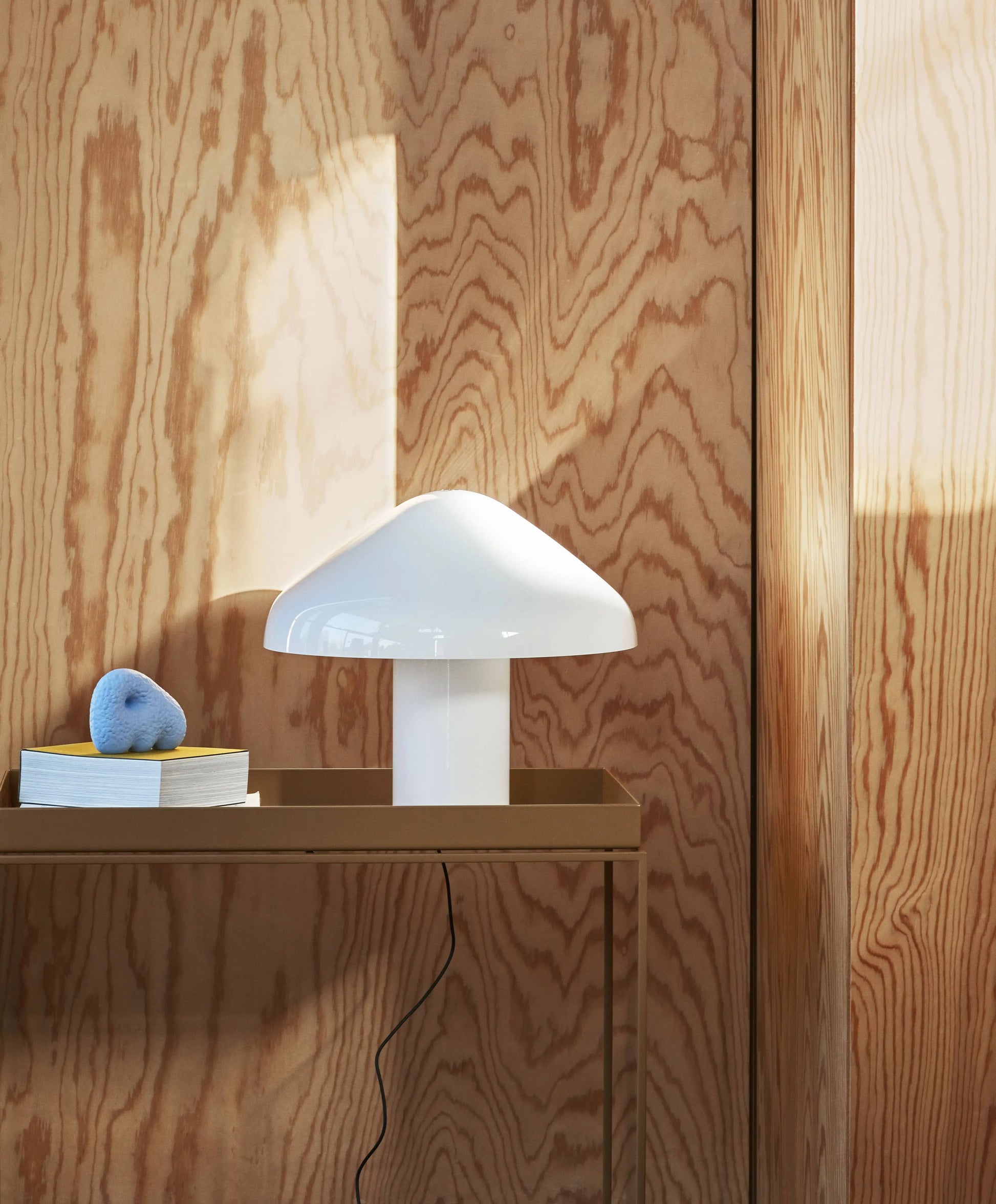 A white HAY Pao Portable Lamp by HAY sits on a brown table against a wooden wall with a stack of books topped by a blue nose-shaped ornament, while sunlight casts a bright rectangle on the wall.
